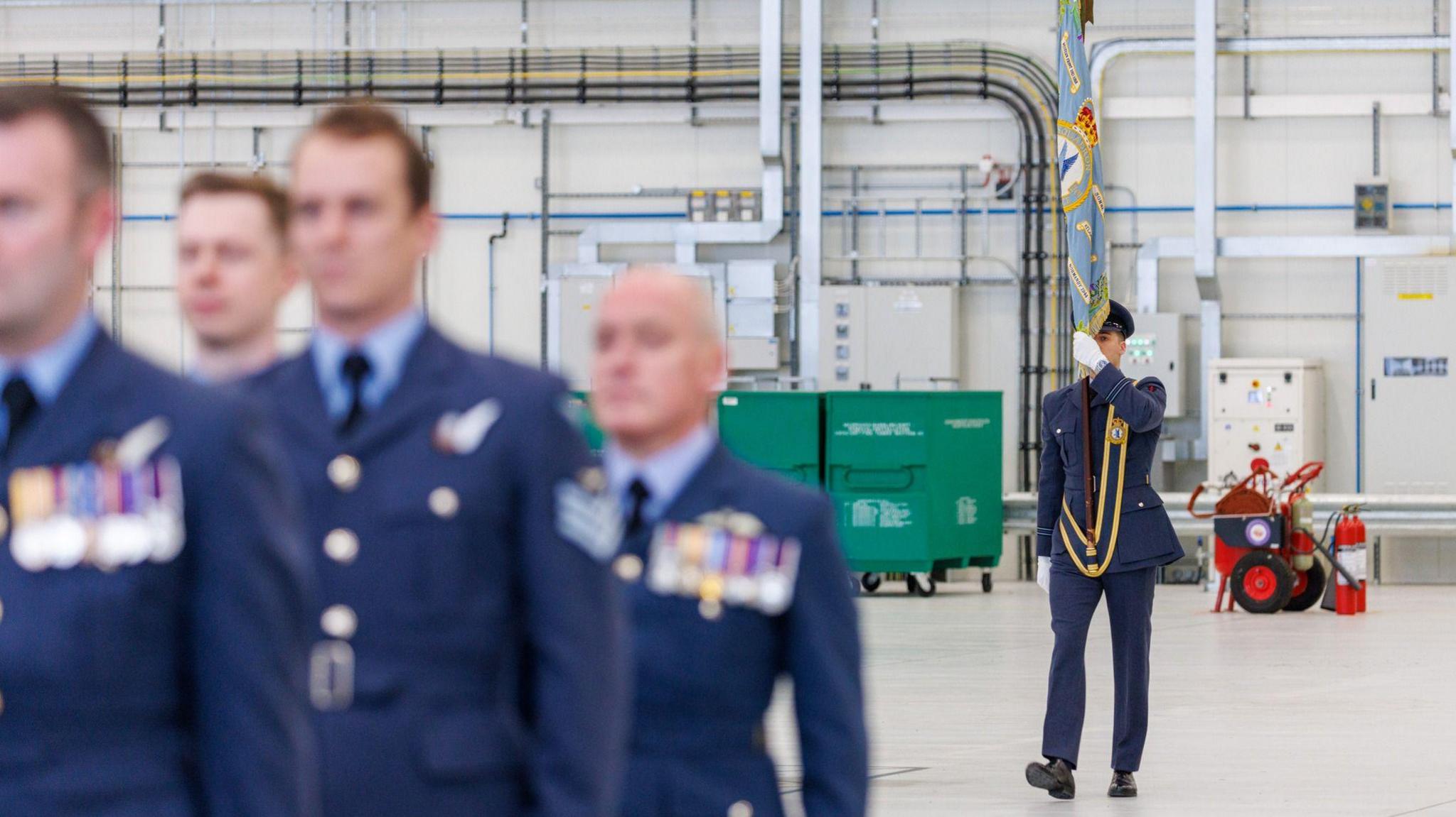 In the foreground, out of focus, stand four men in RAF uniform with medals pinned to their chests and behind them a man in RAF uniform walks towards them holding a standard. They are in an aircraft hangar.