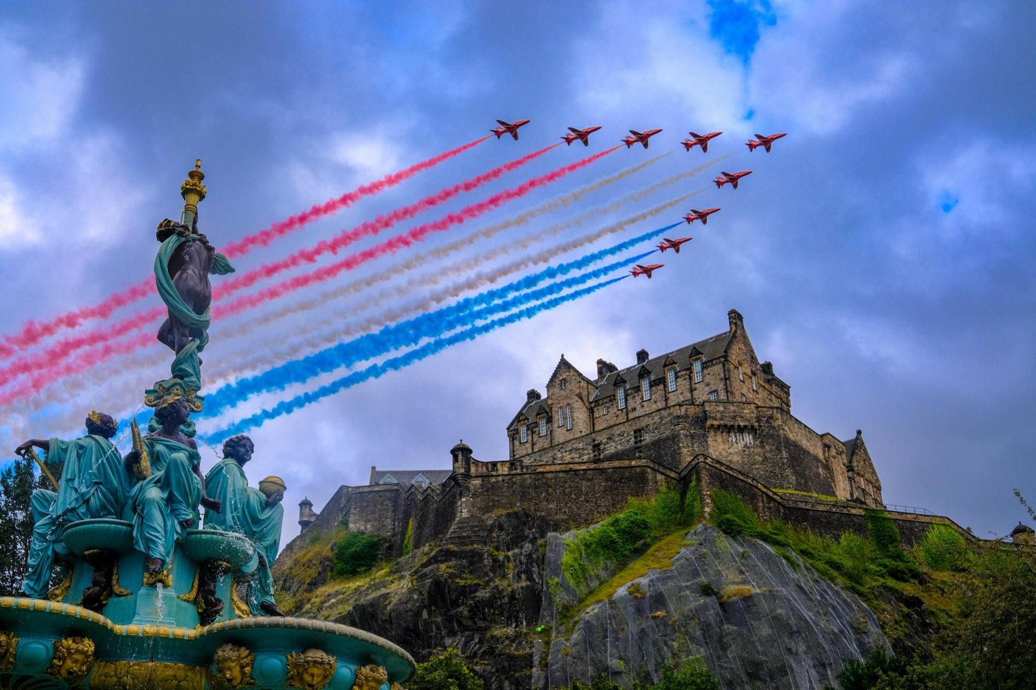 The red arrows flying over Edinburgh castle on an overcast day with the Ross Fountain in Princes St Gardens at the lower left corner. The planes are emitting lines of red, white, and blue smoke as they fly in a V formation.