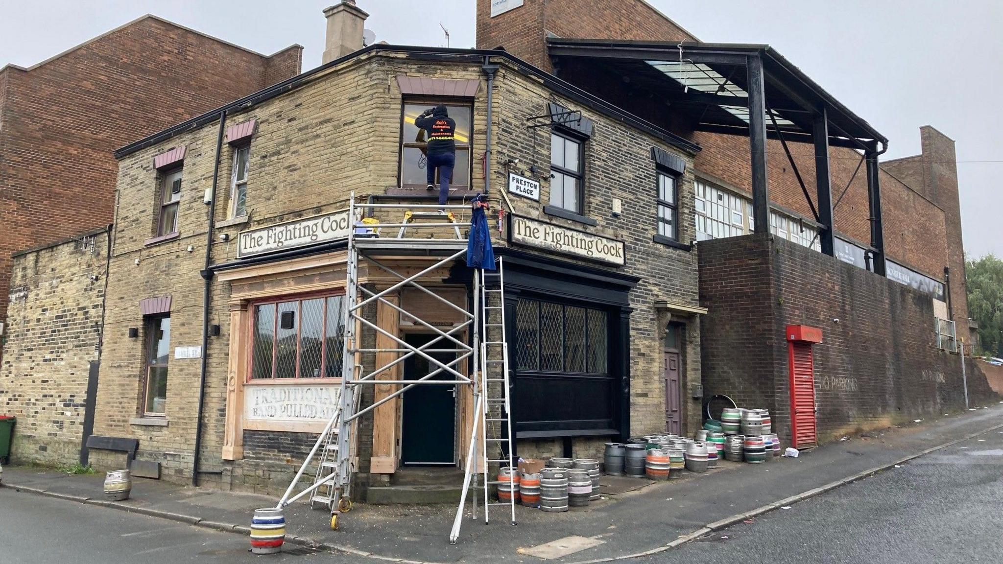 An aluminium scaffolding tower looms over the entrance to the pub which is on the corner of two streets with metal beer barrels outside on the pavement.    