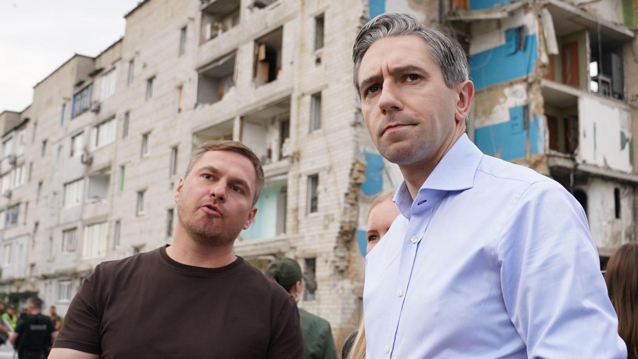 Simon Harris listening to another man speak and frowning. Behind him a badly damaged building