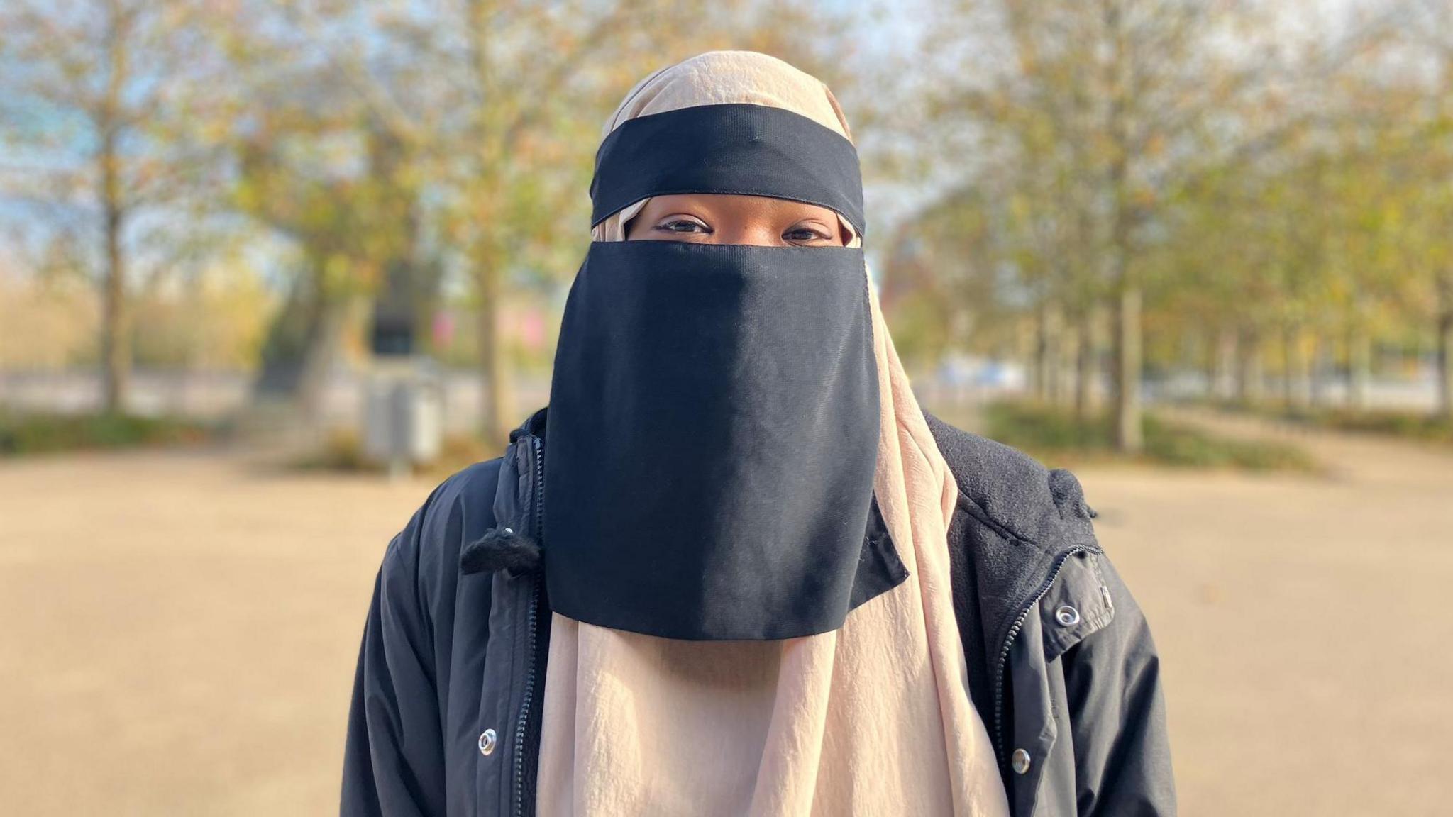 Sahra-Aisha stands in a park on a bright, sunny day with trees behind her, looking directly at the camera. She is wearing a niqab and a black jacket.