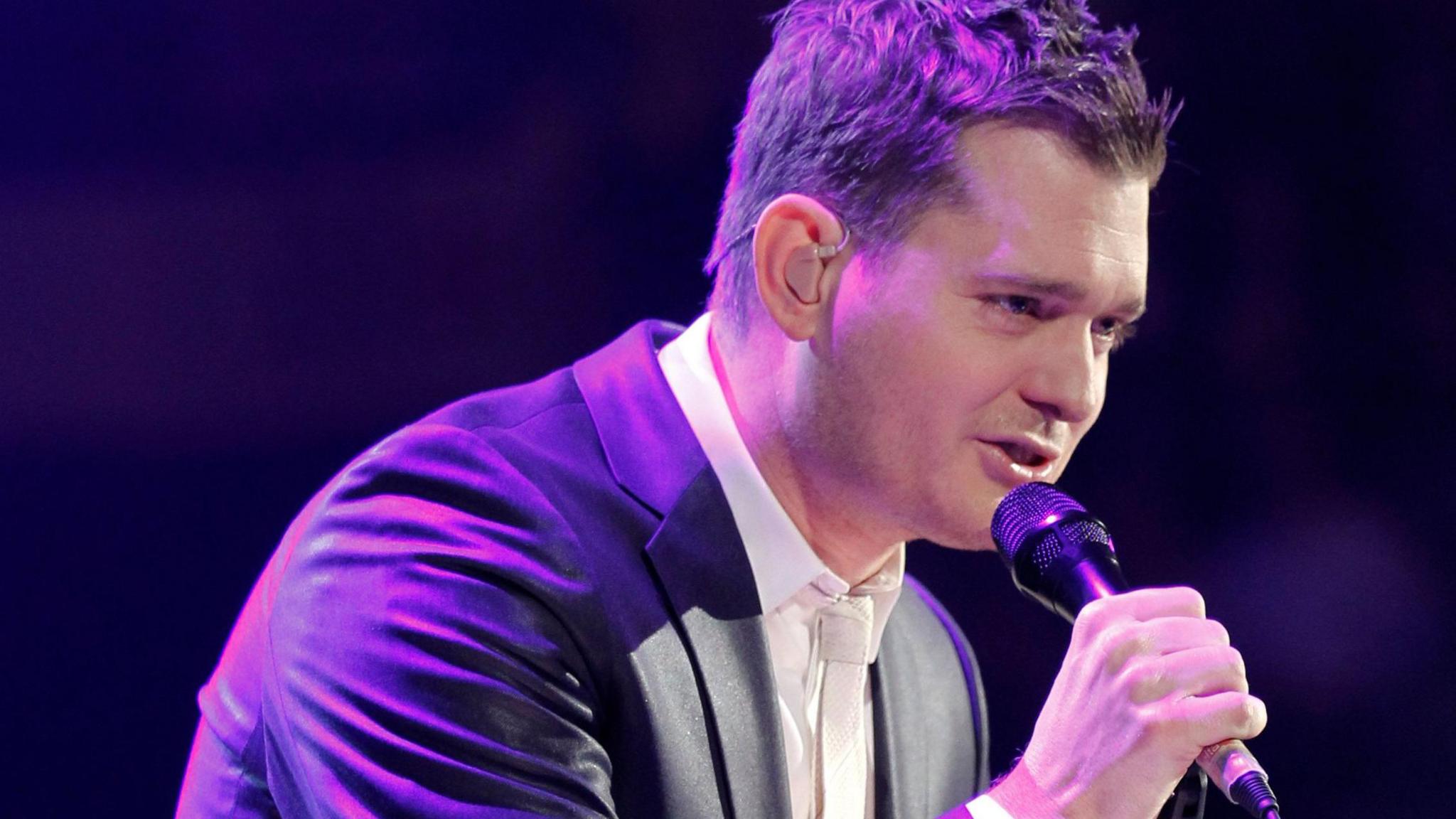 Singer holds microphone and leans forward into a crowd. He is wearing a dark leather jacket and a white shirt and tie. he has dark hair in a modern quiff style