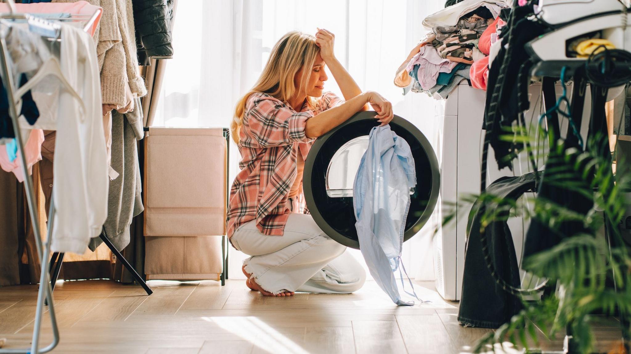 Woman with hand on her head crouches in front of a washing machine with clothes hanging up around her.