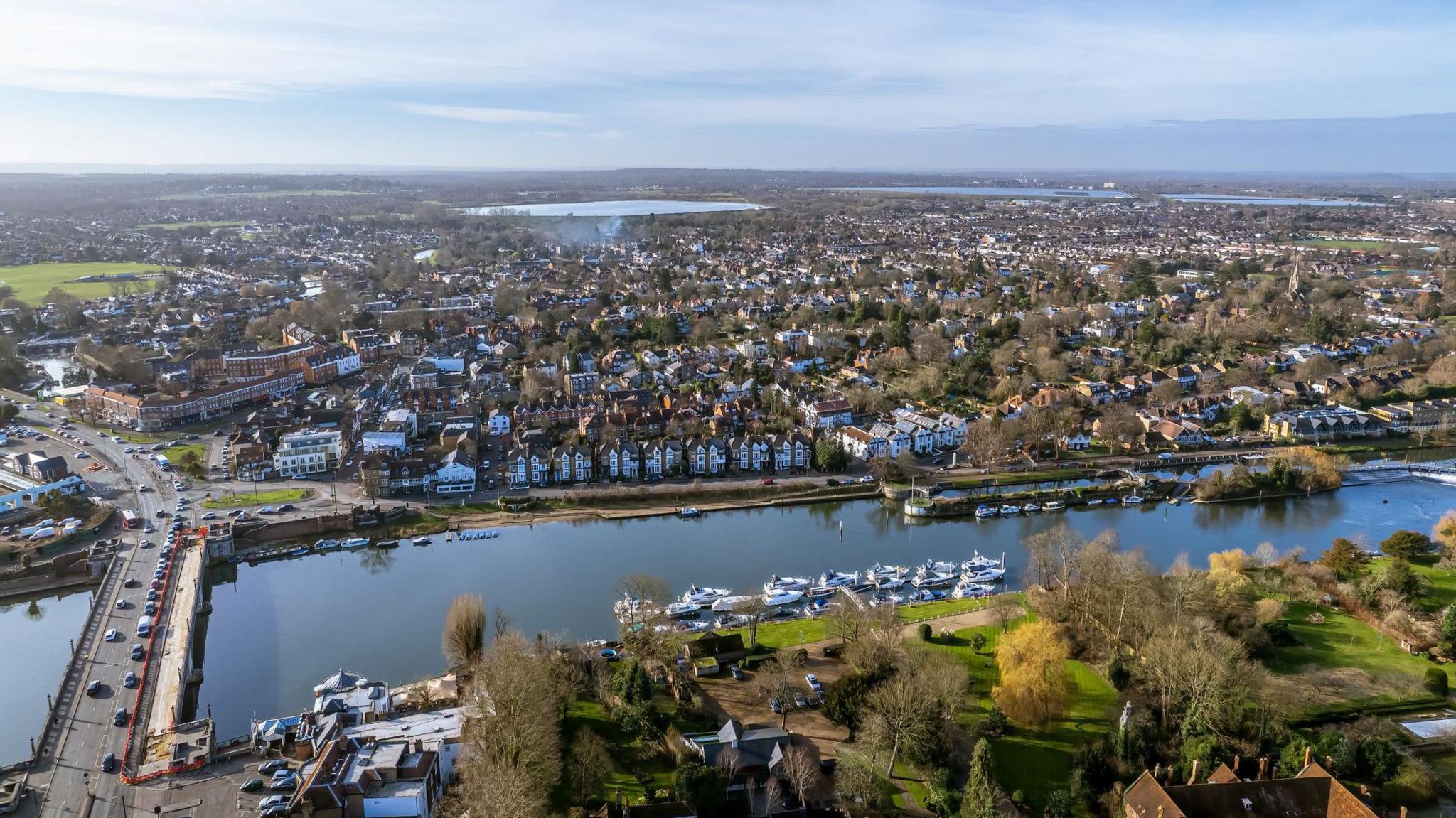 An aerial view of East Molesey which shows the River Thames in the foreground and boats and houses along the edge of the river. Hampton Court bridge is seen in the left of the image and in the distance there are reservoirs and blue sky.