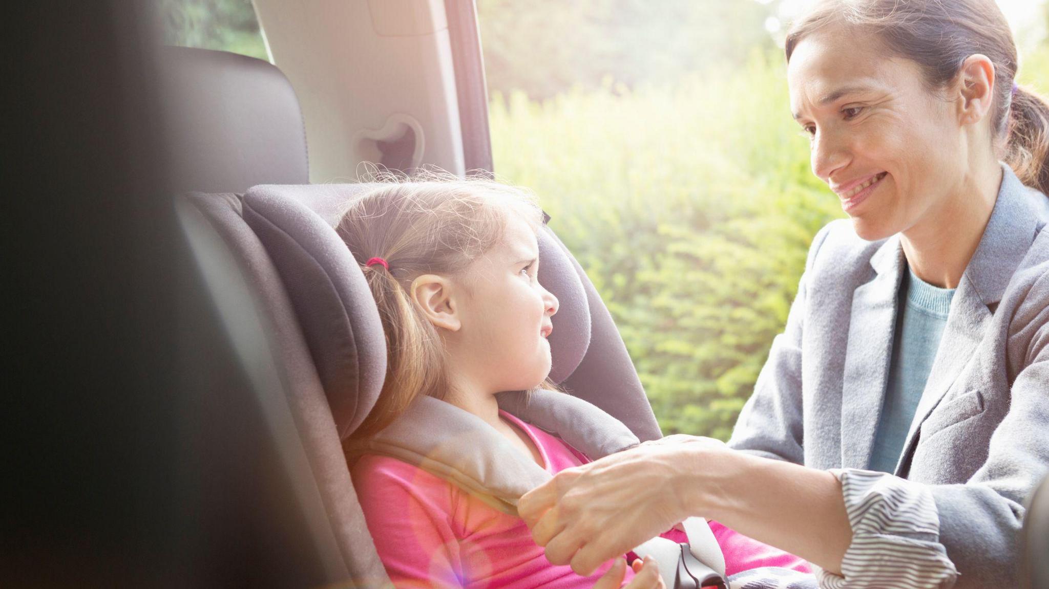 A woman putting a little girl in a car seat. The woman is wearing a grey jacket and blue jumper. She has her hair tied back and is smiling. The little girl is looking at her and wearing a pink top.