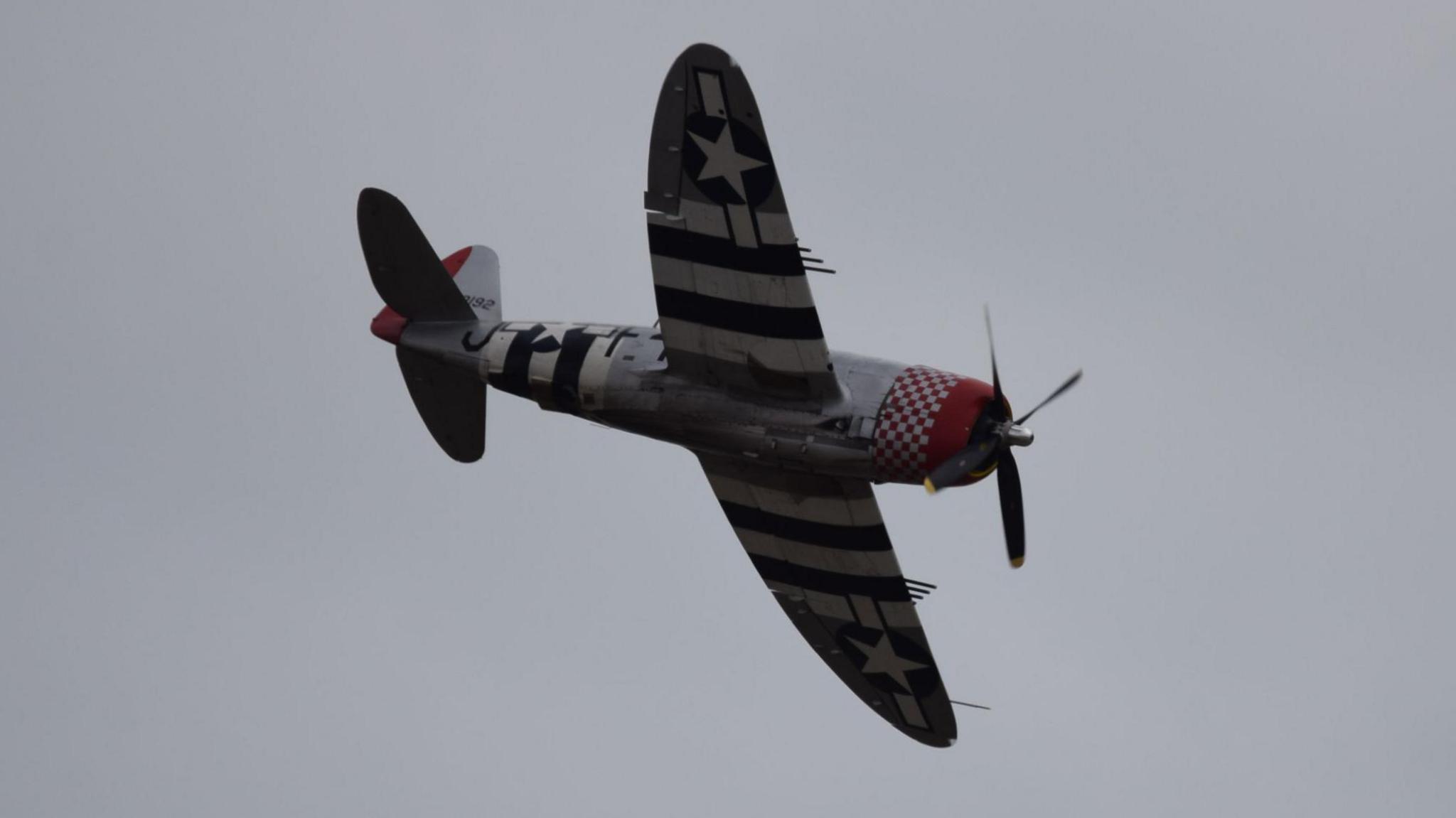 A small plane with the red, white and blue US flag on the underside of its wings. It is flying in front of the backdrop of a grey sky.