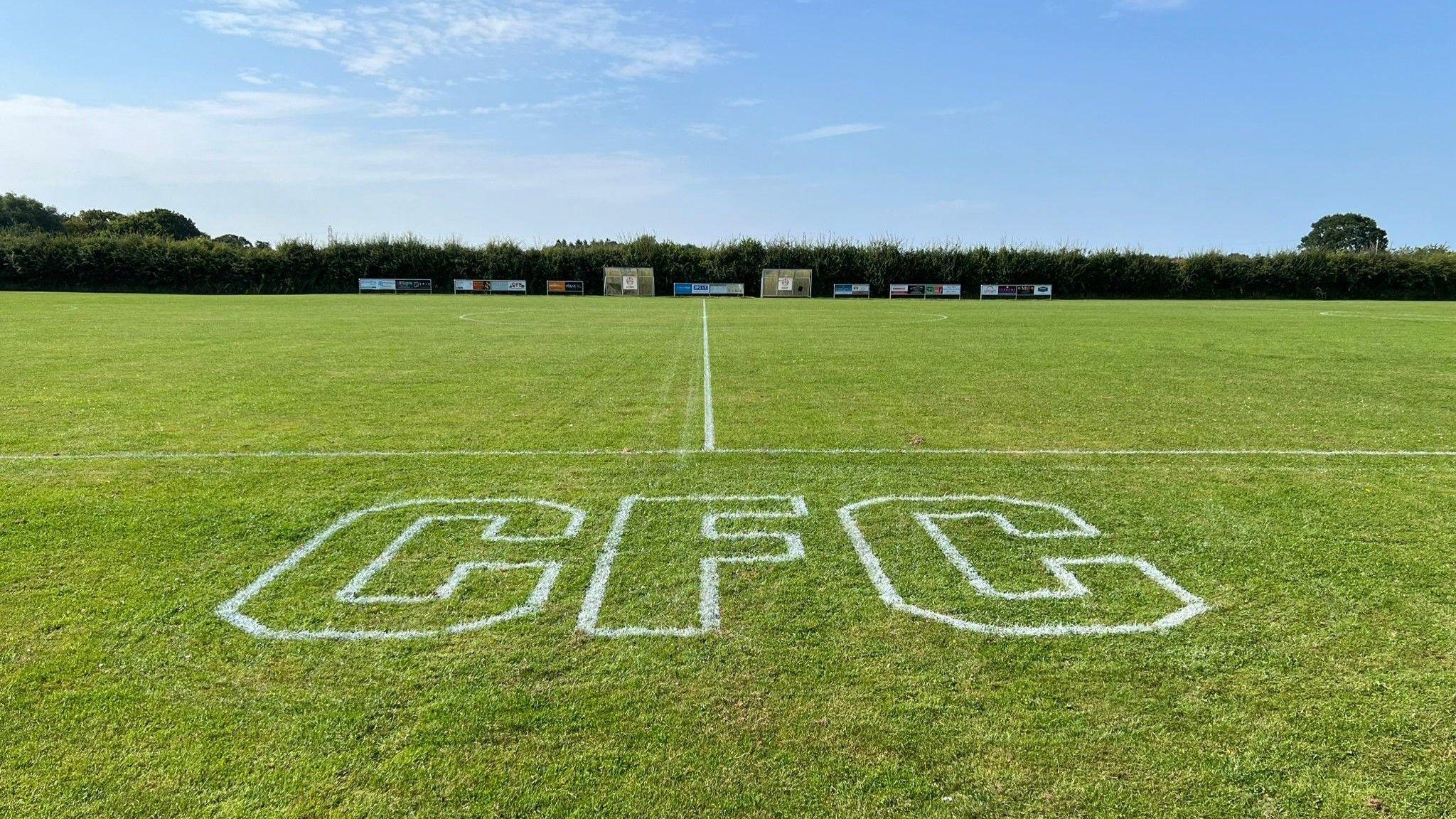 The letters CFC are spelt out in large white paint on the grass next to a football pitch, which be seen in the background lined by a hedgerow.