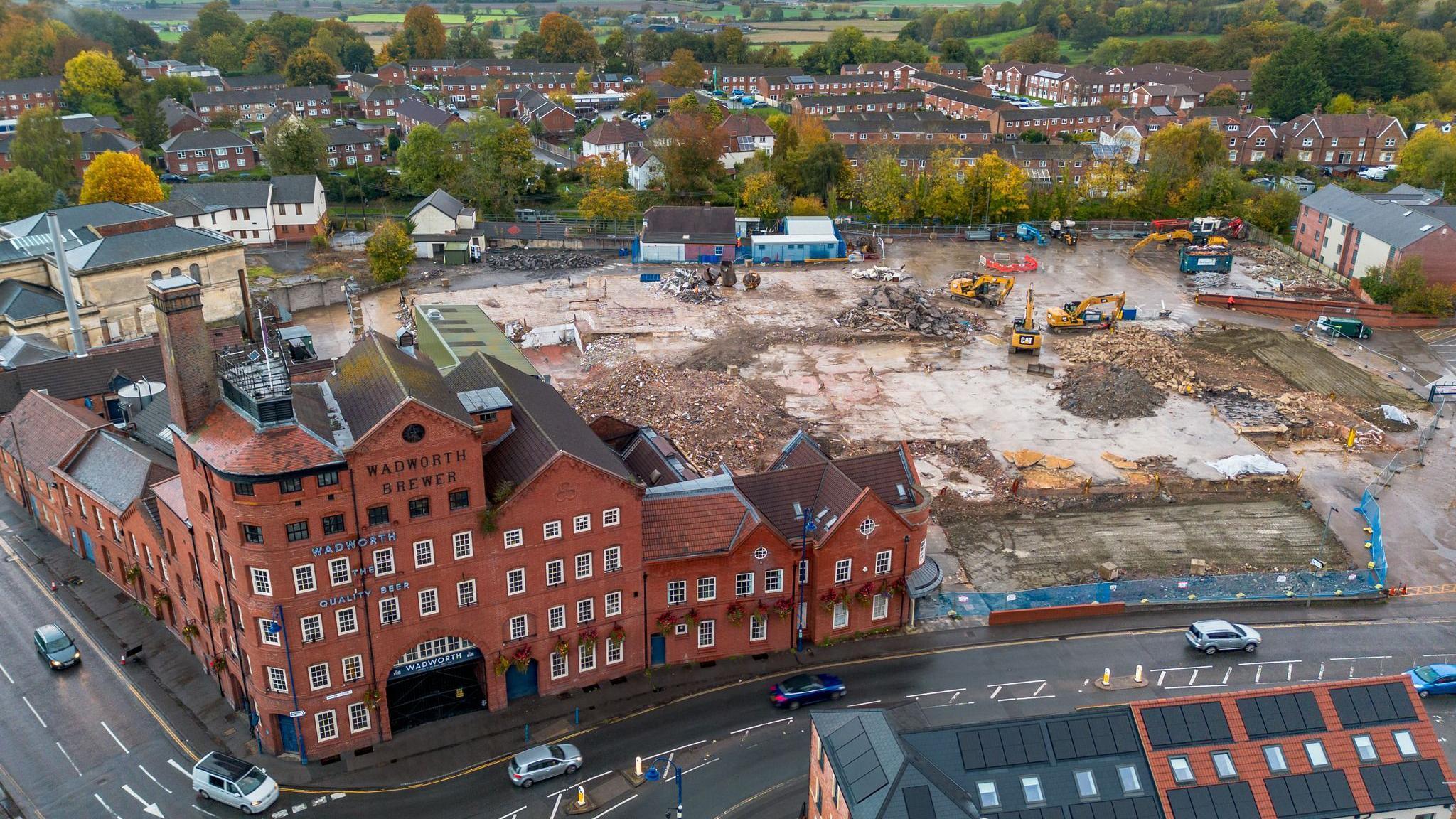 Aerial shot of the site: there is a Victorian red brick brewery building to the left, several storeys high and with a section two storeys high. It has a tower and a large archway with gates. To the right, what now looks like a building site, with big piles of rubble, flattened concrete and big yellow diggers. In the distance, a residential area with trees.