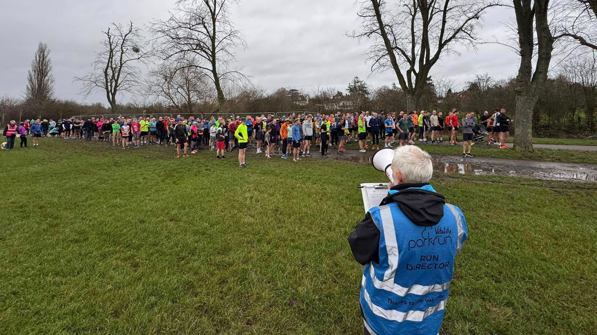 Runners gathered on a muddy patch of grass with a man in blue hi-vis vest that reads "Run Director". Trees without leaves are in the background. 