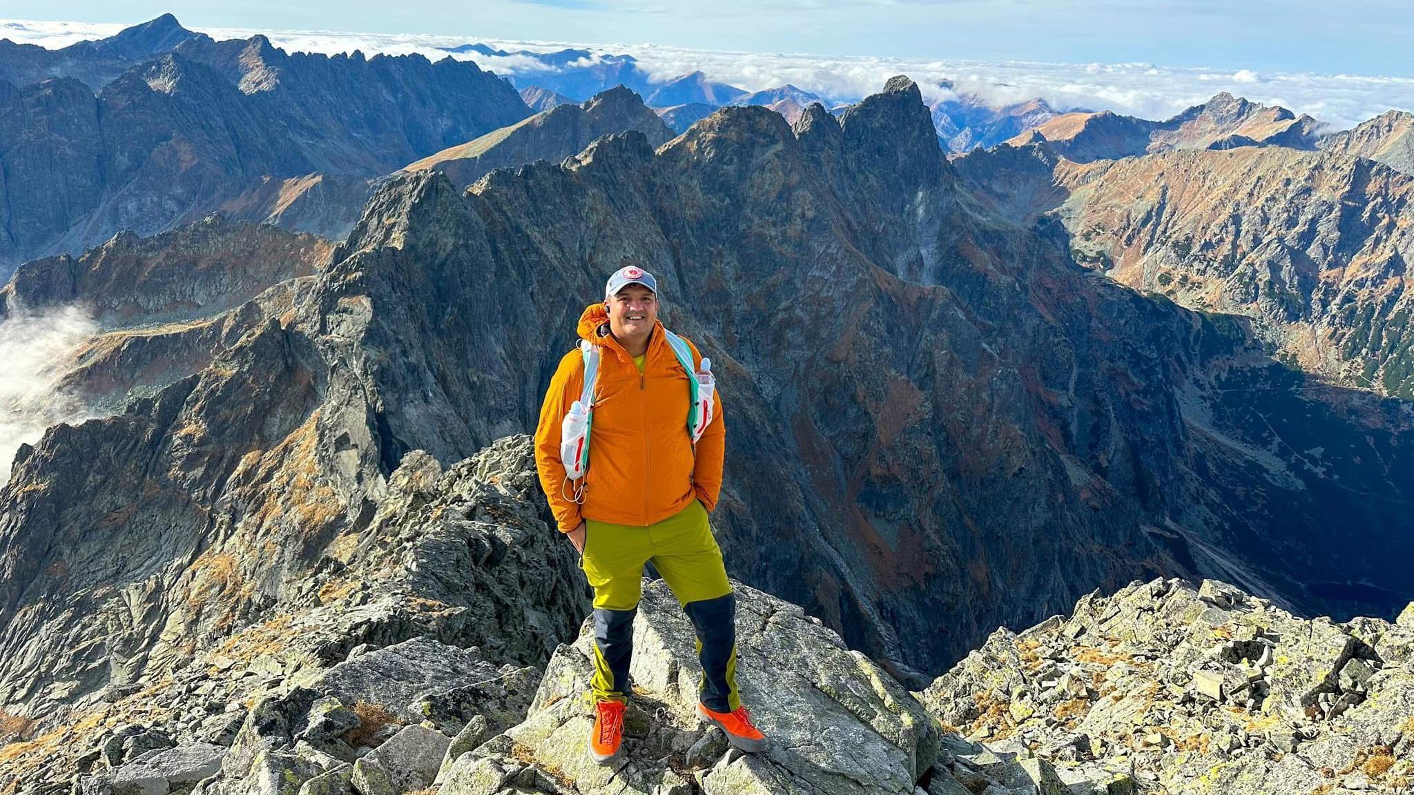 Jay Mistry is wearing a bright orange jumper and lime green walking trousers, he's wearing a cap and smiling at the camera. He is standing at the edge of a large rock with a large mountain range behind him. 