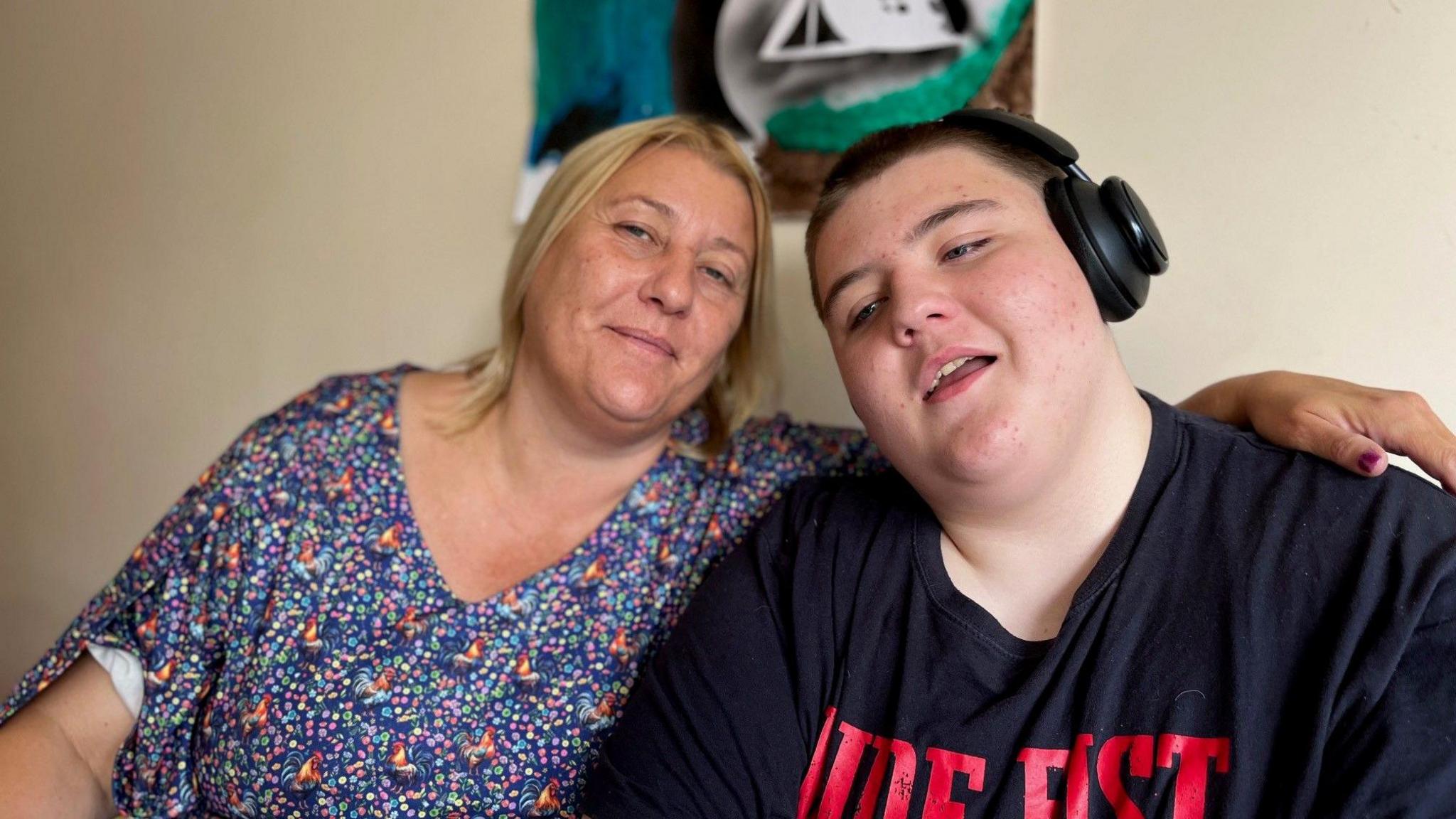 A woman in a blue floral top with her arm around the shoulders of a boy wearing headphones and a black tshirt with red writing on the front