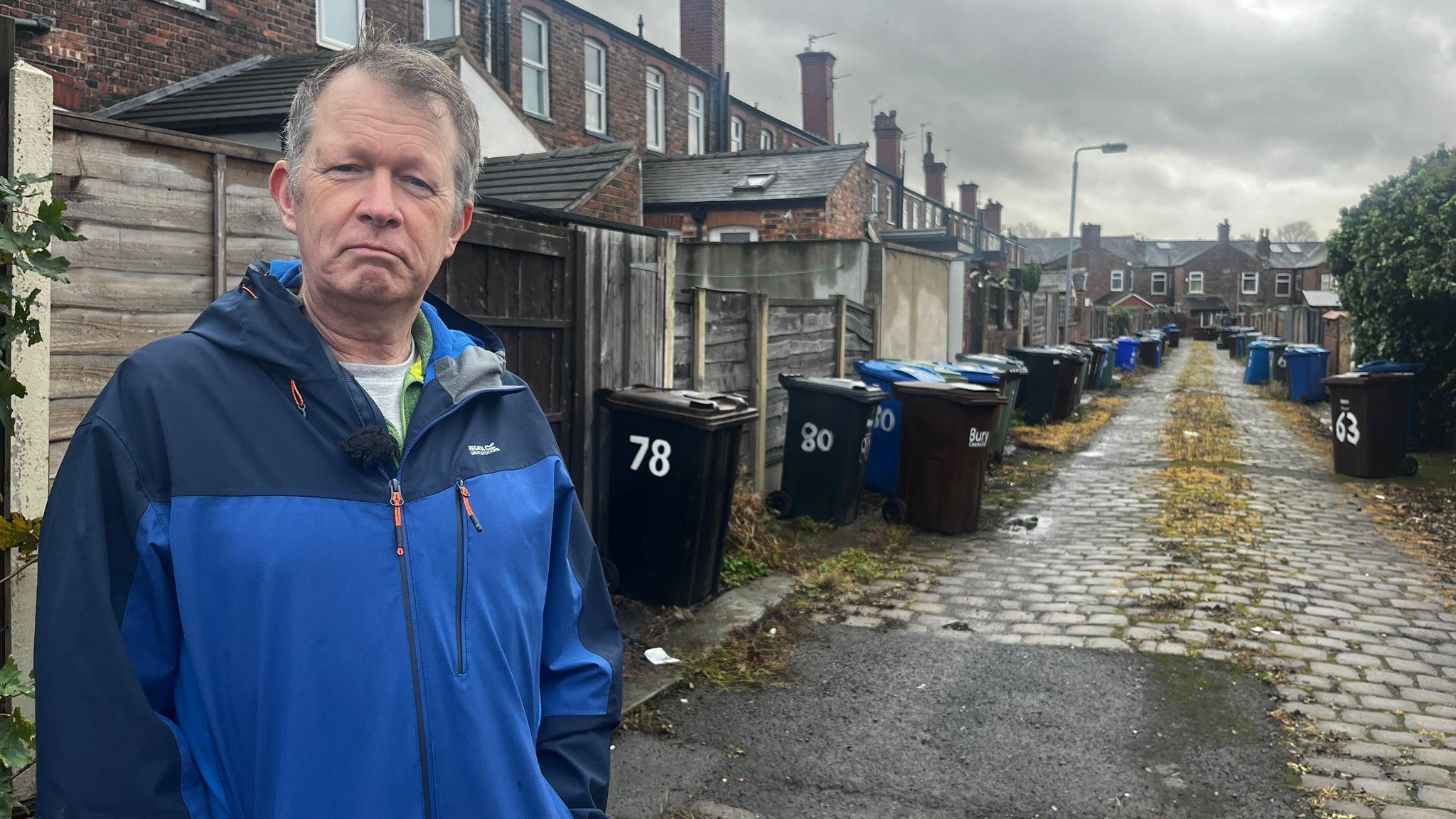 Stuart Atkinson stood in an alley way with lines of wheelie bins the background on a cobbled path