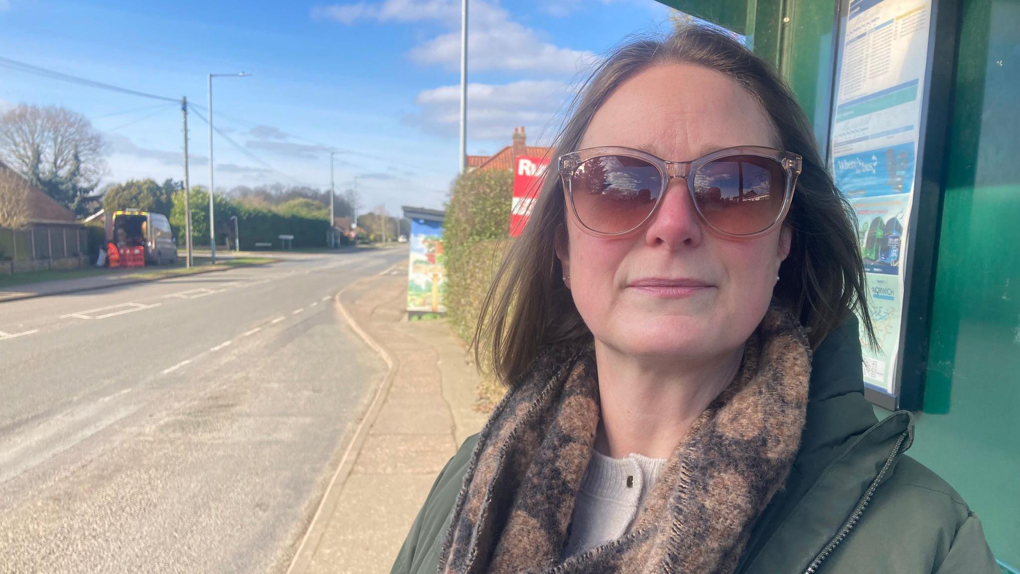 Woman in dark glasses standing at a bus stop. She is wearing a green coat. There is a road beside her. 