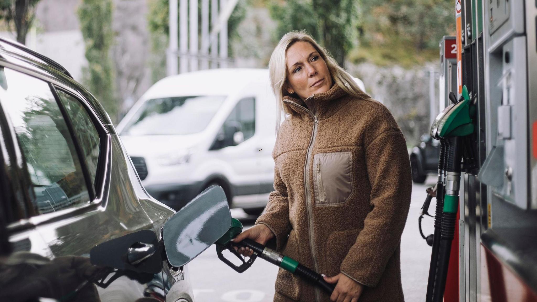 A stock photo of lady in a brown jacket filling up her car with fuel at a petrol station.