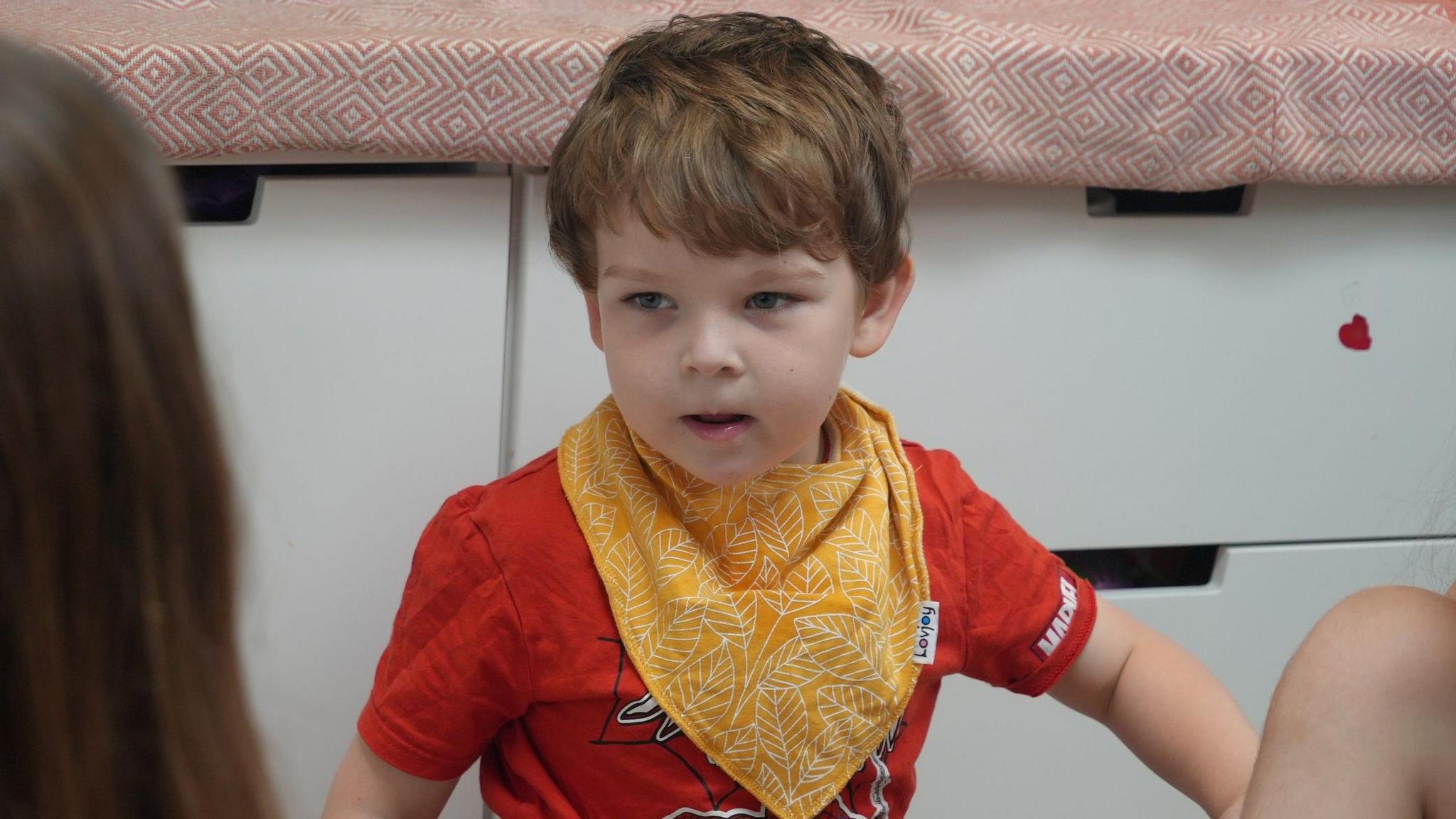 A close up of Bobby looking at one of his sisters. He wears an orange neckerchief with white leaves on over a red T-Shirt.