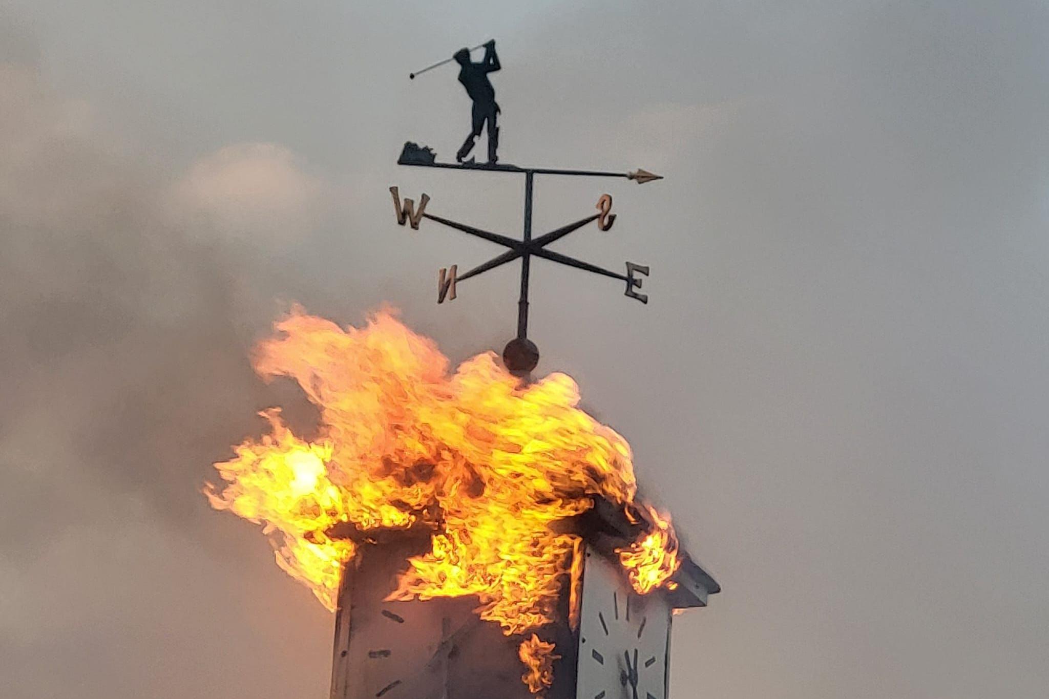 Rood alight with square clock tower with weathervane golfer silhouette on the top - flames are coming out from the sides of the clock.