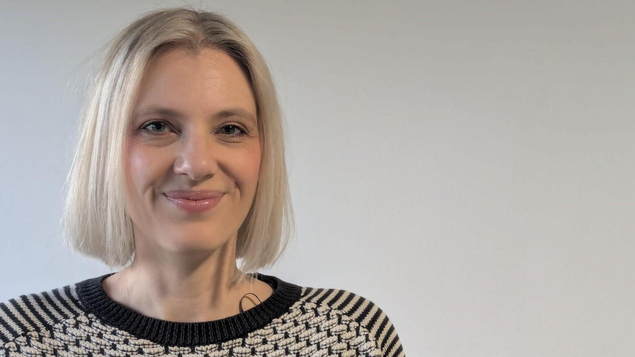 Head and shoulders of a woman with shoulder length short blonde hair, standing in front of a white backdrop.