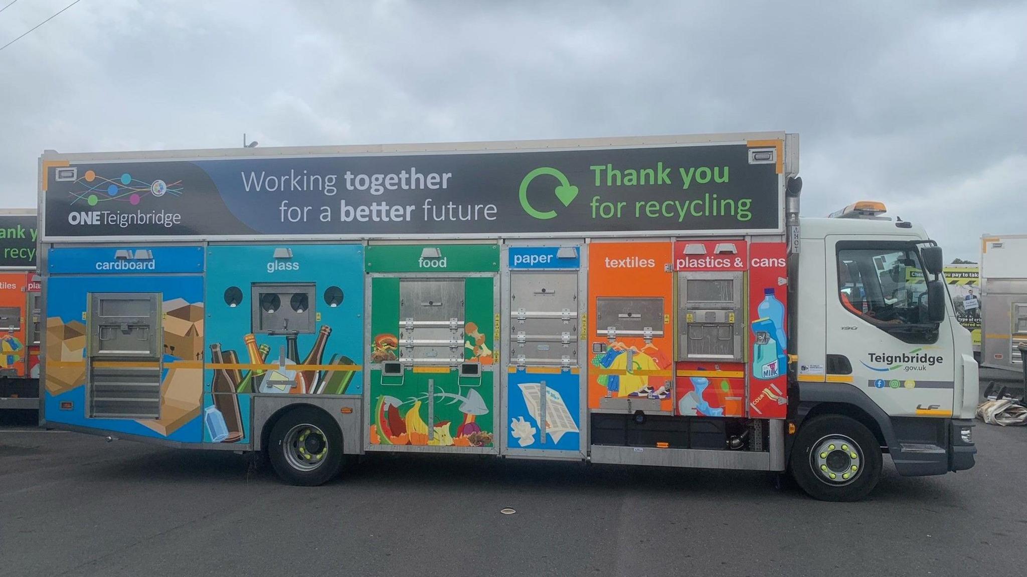 A recycling collection vehicle. It has various compartments for recycling. It's covered in imagery representing the sort of items people can recycle, like food and plastics