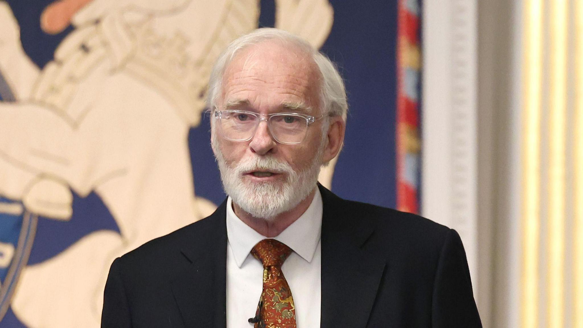 Actor Ian McElhinney with grey hair and a grey beard. He is wearing a black suit, white shirt, orange tie and glasses with a clear frame. 