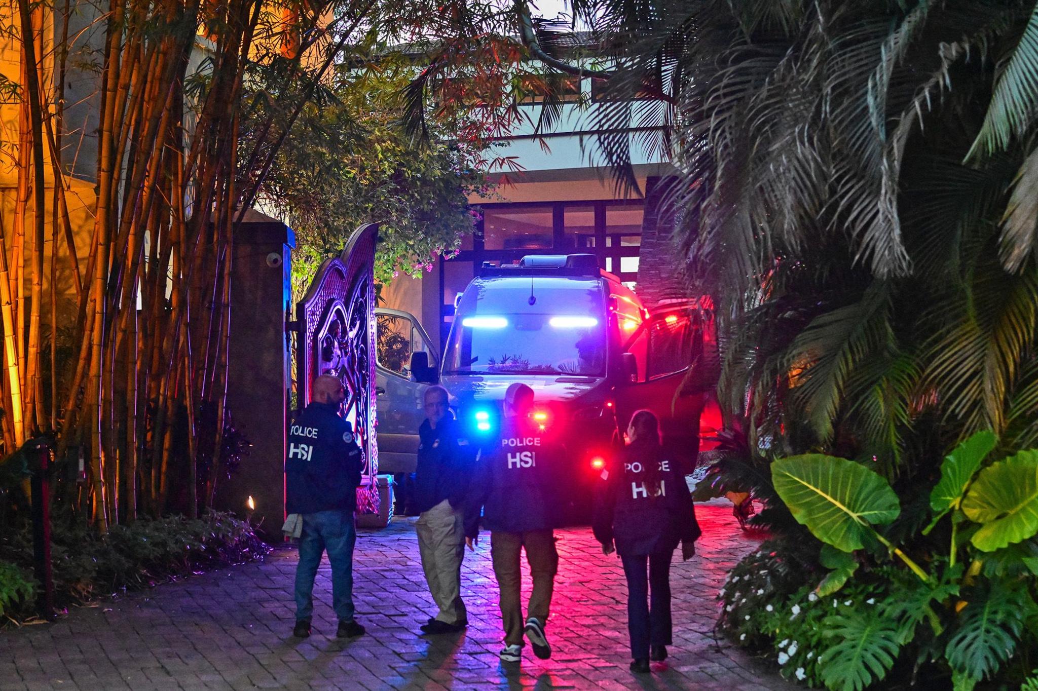 Police HSI officers walking toward a building at night, with a police vehicle flashing lights nearby, surrounded by greenery and illuminated by various lights.