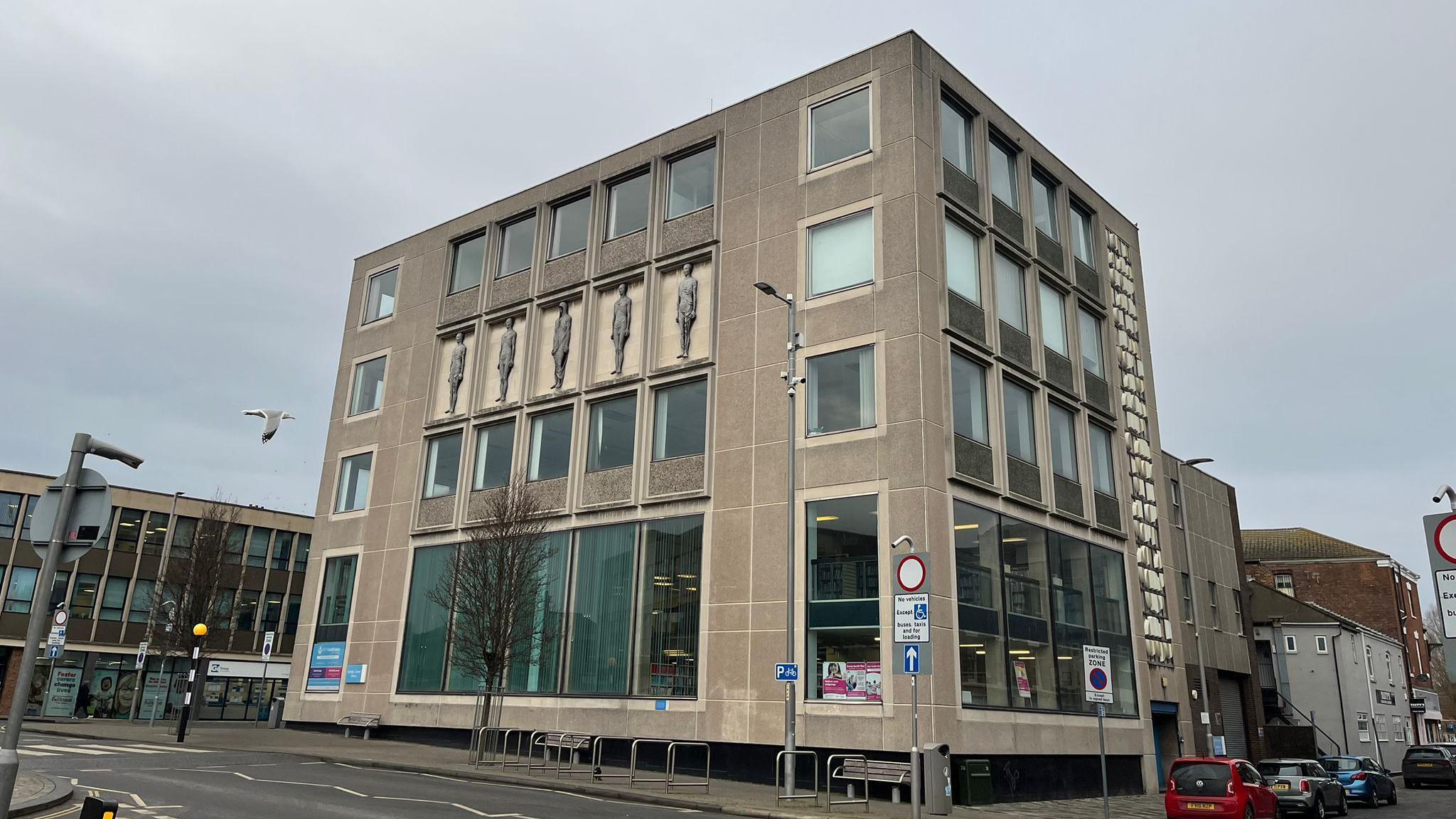 The outside of Grimsby Central Library, which has several rectangular windows and light brown stone on the outside. There is a design on the front of five people standing upright. It is  next to a road and near other town centre buildings.