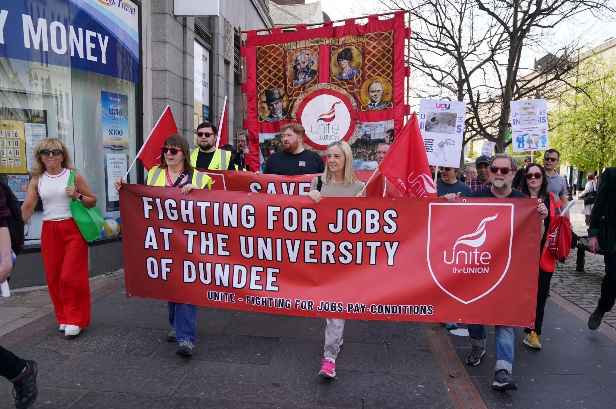 Protestors carrying flags and marching and holding up a bright red banner which has fighting for jobs at the University of Dundee written on it in white letters. 