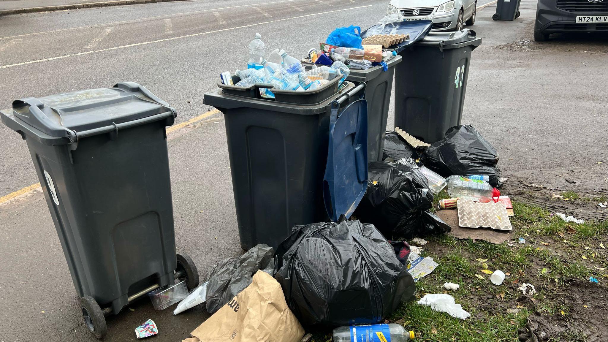 Image shows four dark grey wheelie bins on a street, two have their lids open and are overflowing with recycling. There are also black rubbish bags on the floor with waste spilling out onto the road and grass verge.