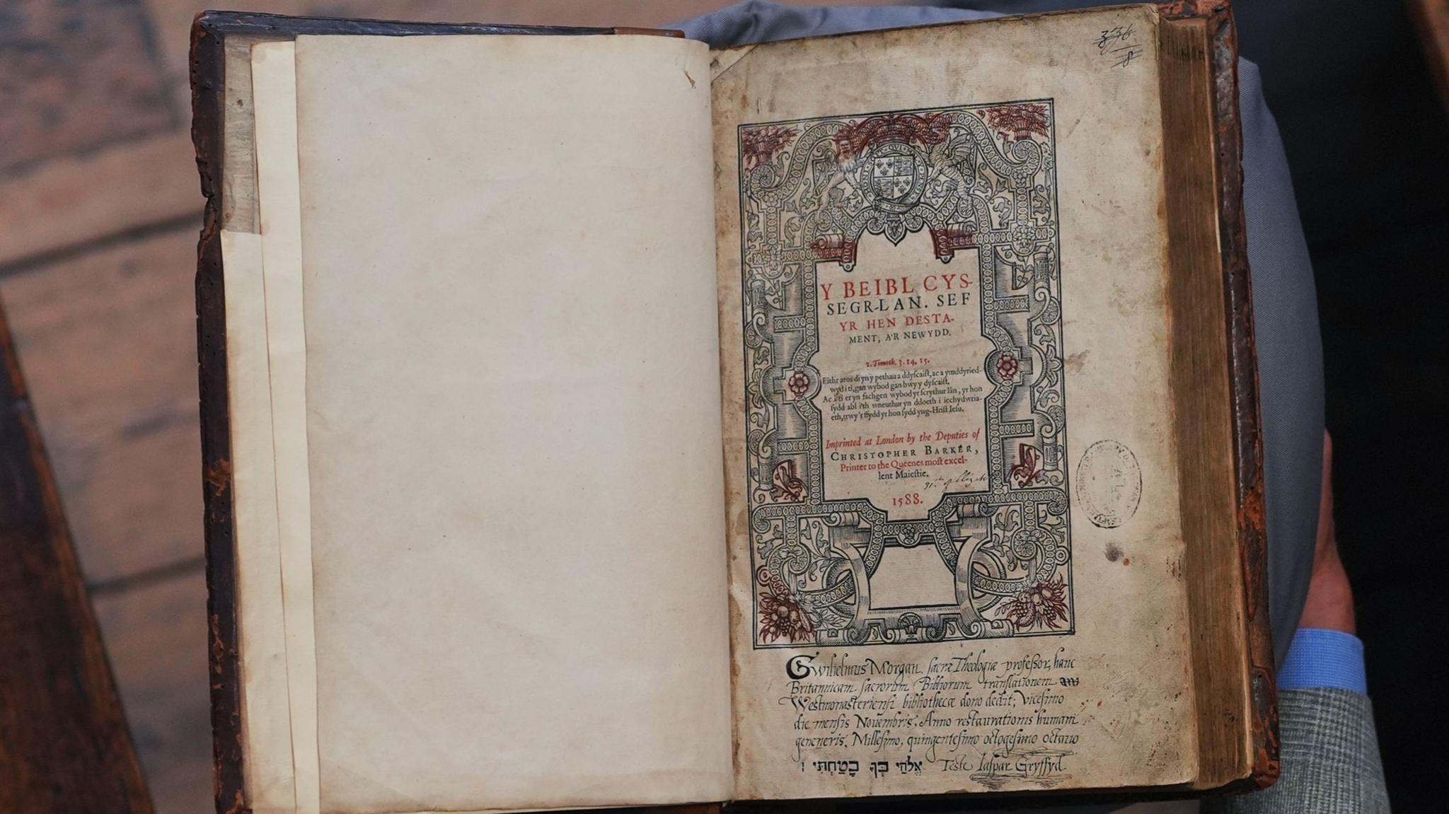 A man in a grey suit with grey hair holds an antique-looking book. The photo is captured from above so the viewer looks down on the scene. The man is stood on a wooden floor with a red carpet laid down. The book has a beautiful Celtic design on the right hand page and calligraphy. The paper in the book is yellowed and spotted with age.