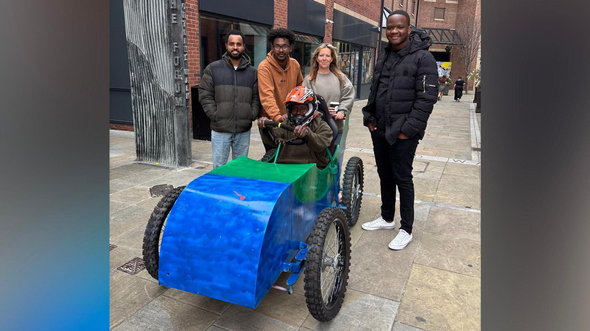 A person in a red helmet sits in the green and blue cart, surrounded by four other people who have smiles on their faces.