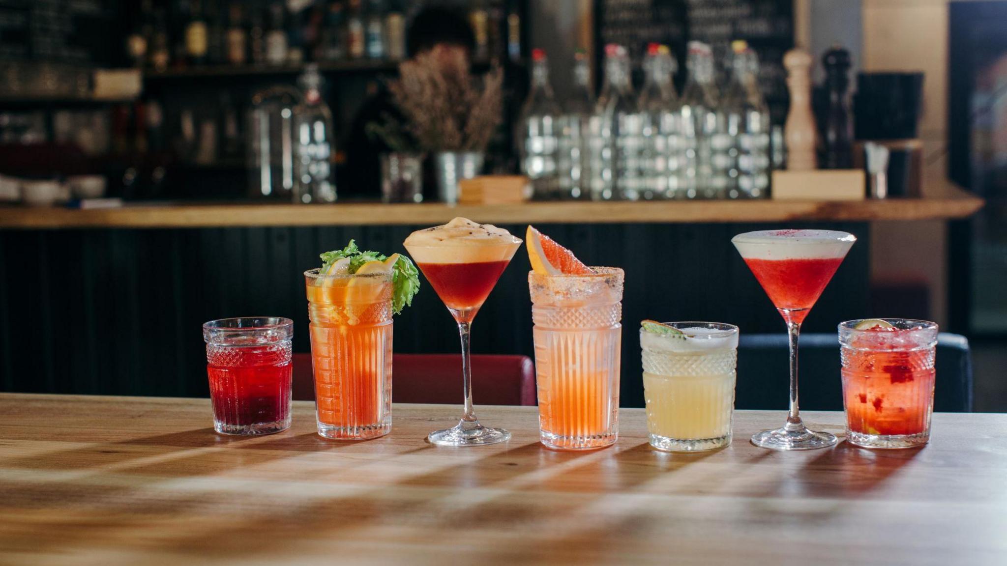 Several colourful cocktails on a table in a bar. Some are in martini glasses, some are in tumblers and some are in highball glasses.