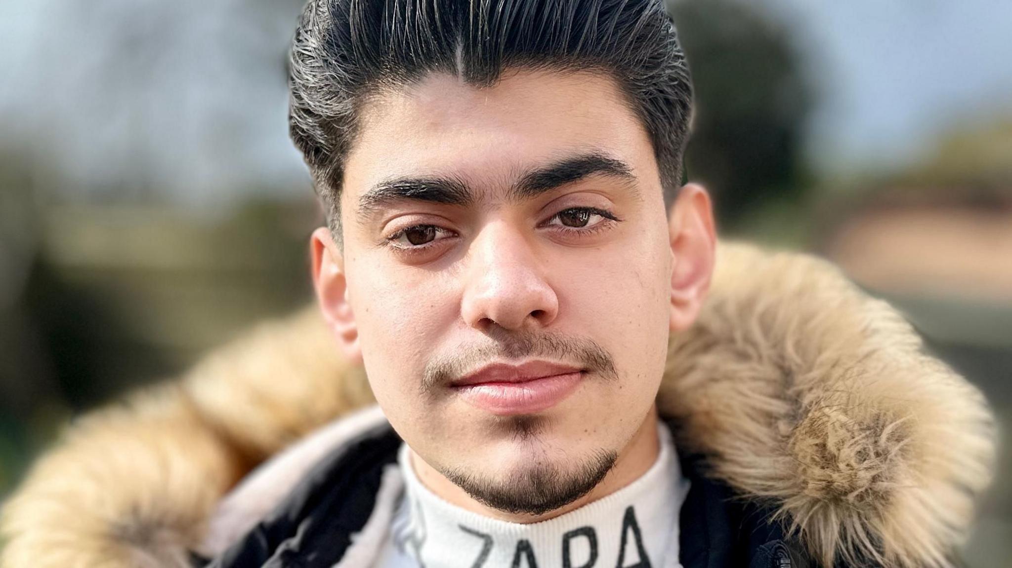 Portrait photo of Ahmad Othman, who has dark hair, brown eyes and a neat goatee beard, looking to camera. He is wearing a coat with a furry hood and a while roll-neck top.