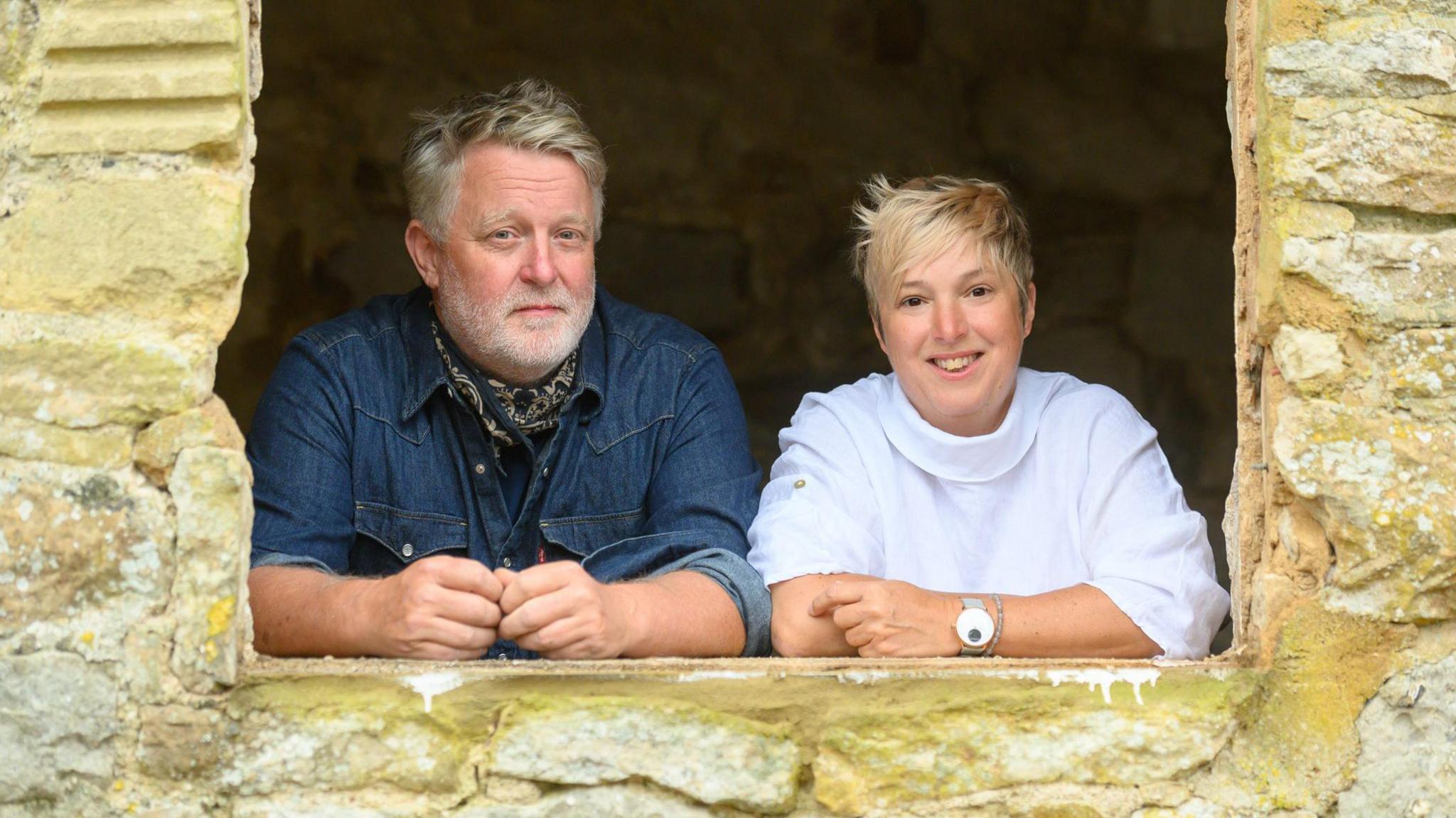 Mark and Jules leaning through an open window in a light washed brick building. Jules is on the right, with short blonde hair and wearing a white shirt and watch. Mark is on the left, wearing a blue denim shirt rolled up at the sleeves and a dark bandana around his neck. He has dark grey hair swept to the side and a full white beard. They are both smiling at the camera.