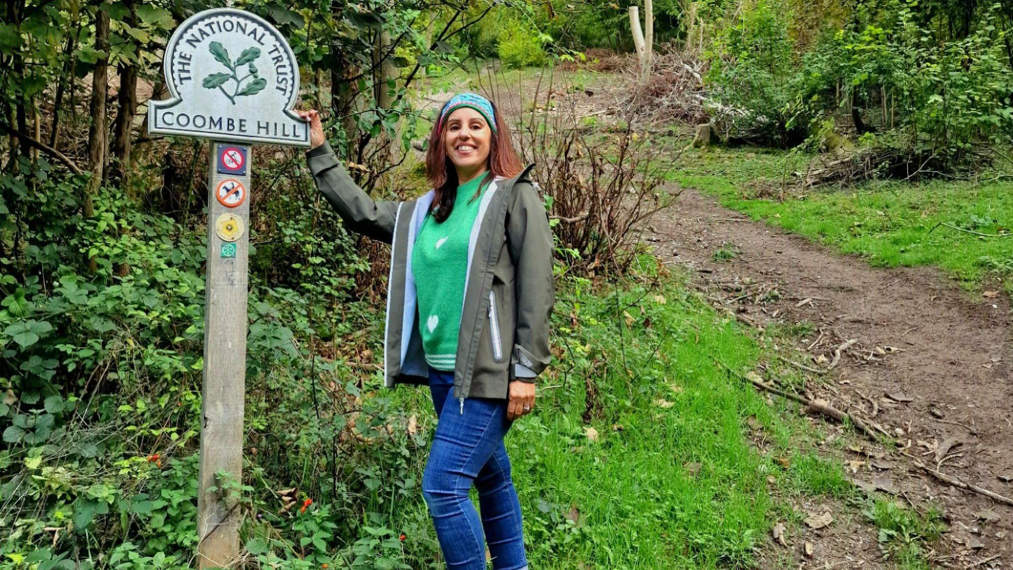 Geeta standing next to a National Trust sign with its logo and the words "Coombe Hill". It is in woodland and she is holding onto the right side of the sign. She wears blue jeans, a bright green jumper, a sage gren jacket and a turquoise patterned headband. There is a dirt track behind her.