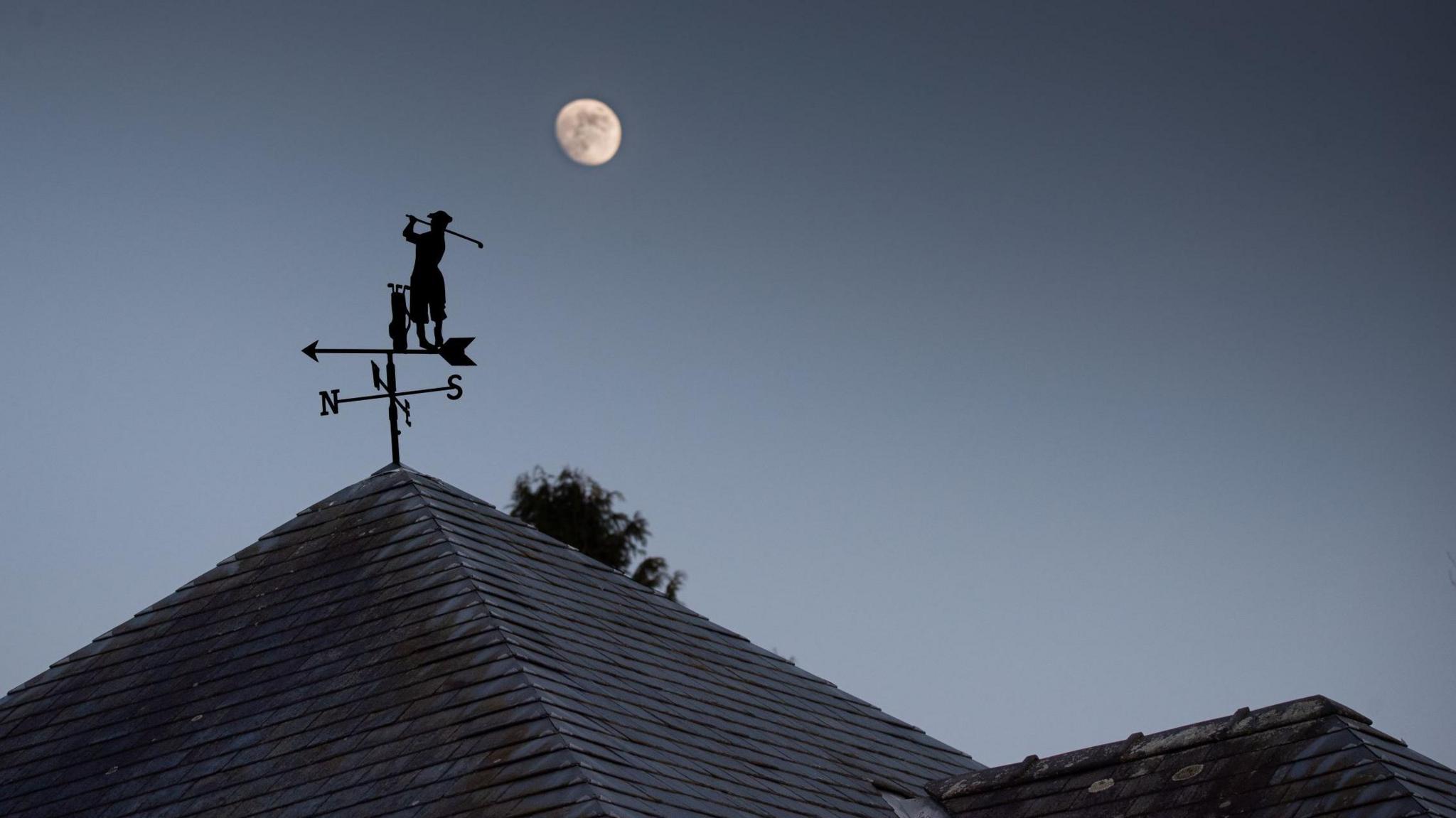 A weather vane on the roof of a house has a silhouette of a golfer hitting a shot. Just above the figure is the moon. The sky is dark and starless.