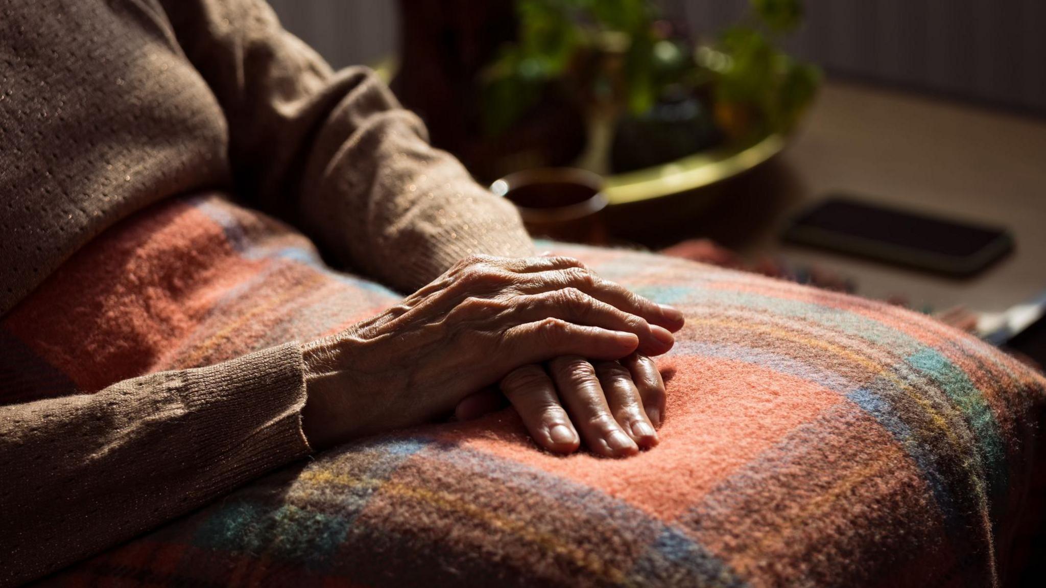 An older person is sitting in a chair with one hand placed on top of another. They have a tartan blanket on their lap.
