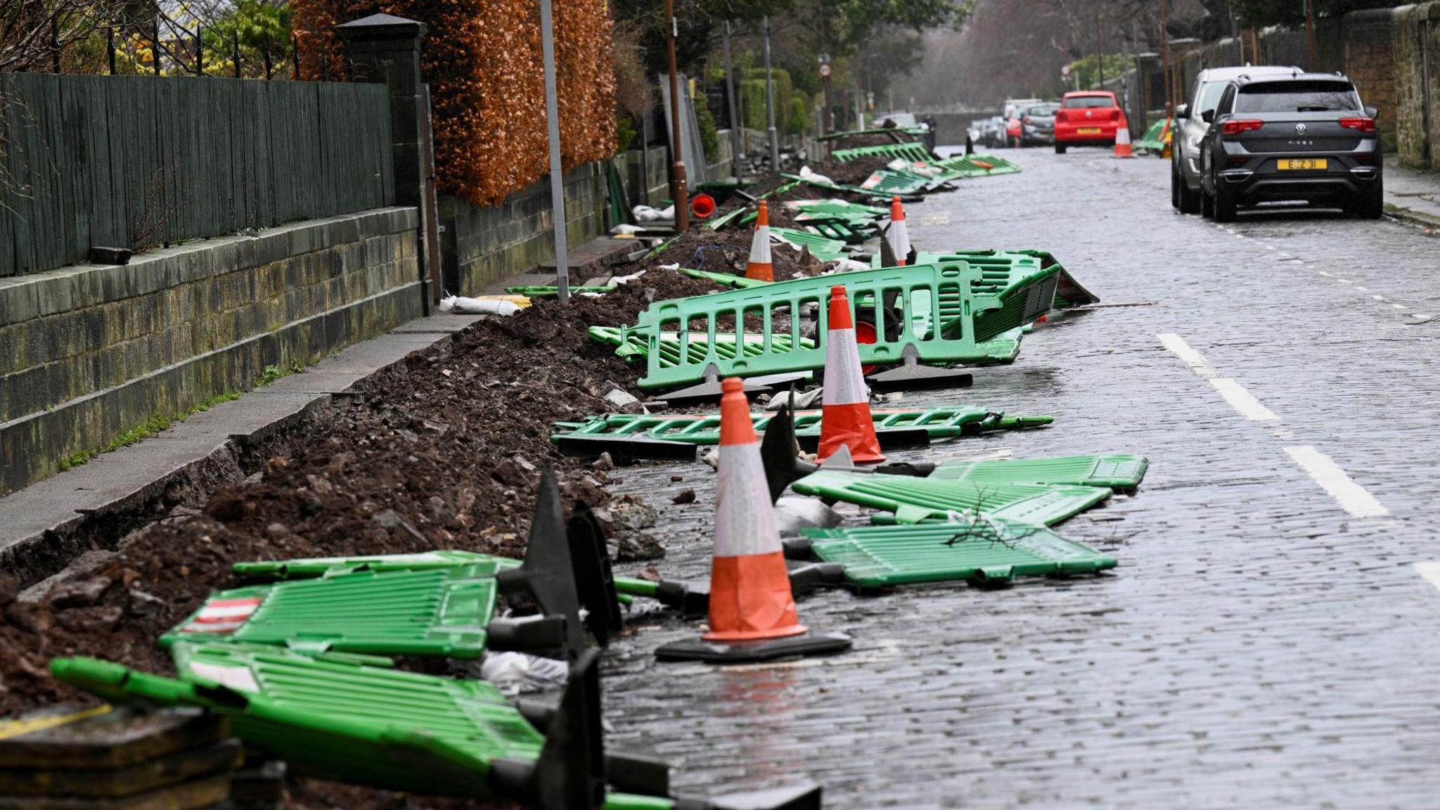 A residential street in Edinburgh with overturned roadwork barriers and traffic cones on the left and cars parked on the right