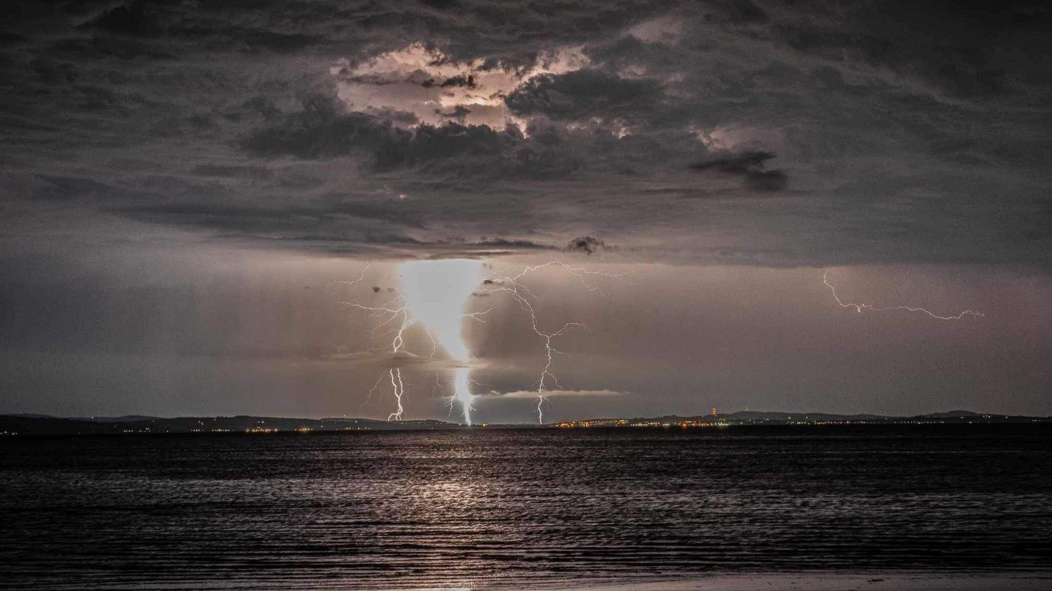 Lightning pictured in the distance coming from a grey cloudy sky. The sea is at the bottom of the photo.