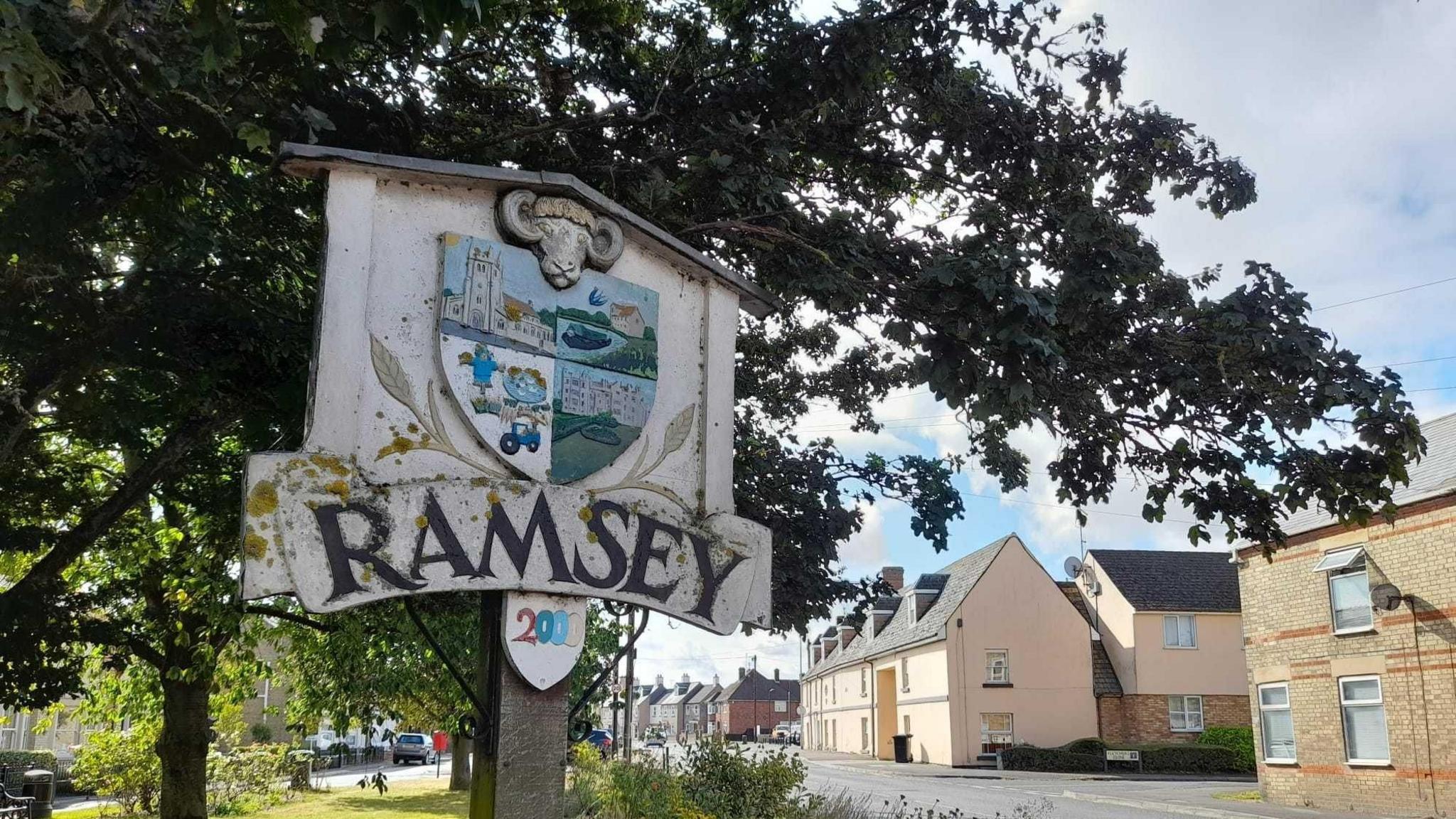 A white town sign which says Ramsey in black letters. Above the lettering is a shield which is quartered to show four scenes from Ramsey, including the parish church, a tractor, a stately home and a river. Above that is a ram's head. Behind it are trees in leaf and to its right are houses and a road