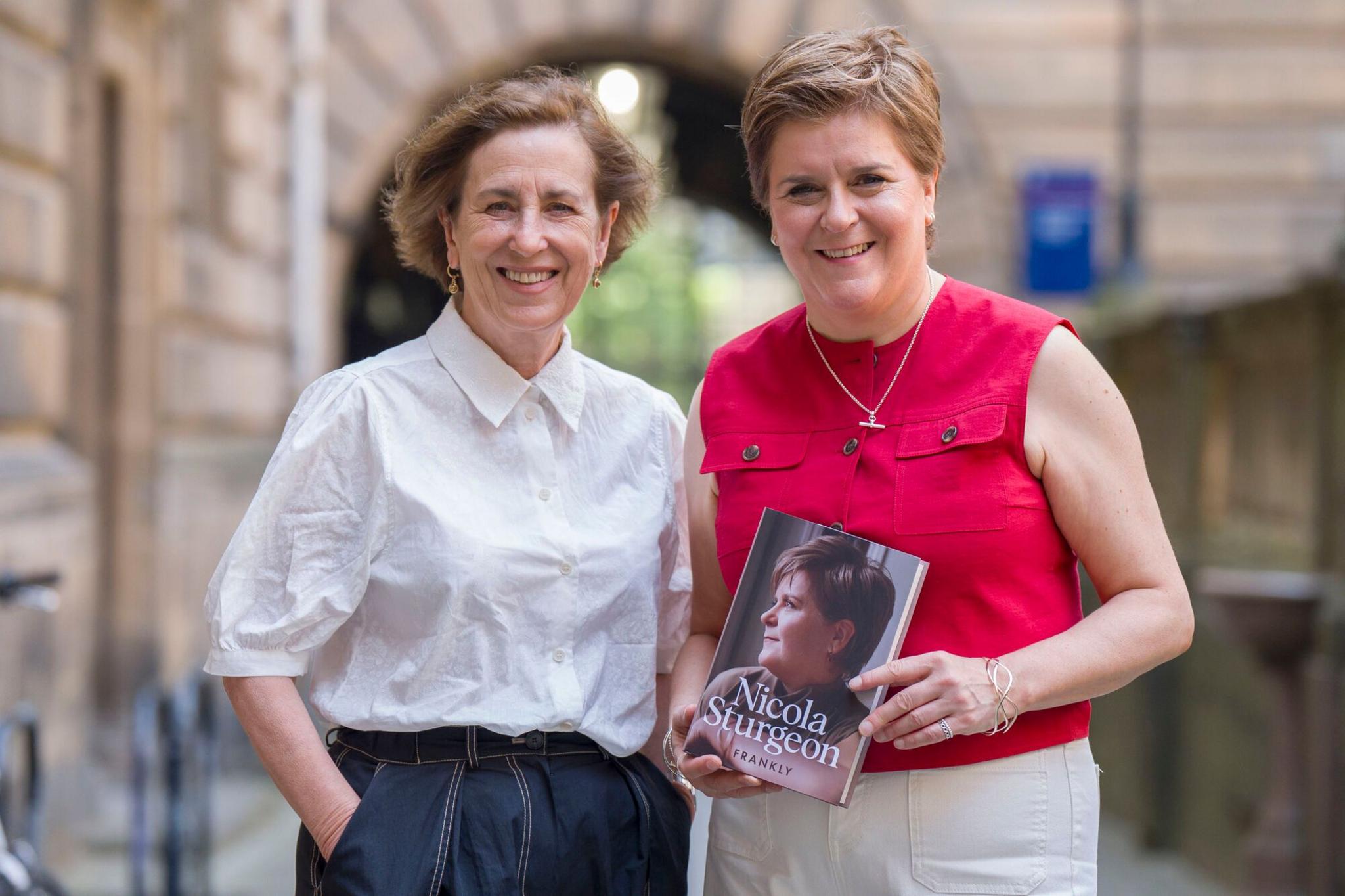 Kirsty Wark has short brown hair and smiles at the camera. She wears a white shirt and dark trousers. She stands next to Nicola Sturgeon who smiles at the camera. She has short fair hair and wears a red top and white trousers and holds a copy of her book which has her face profile pictured and is called Frankly