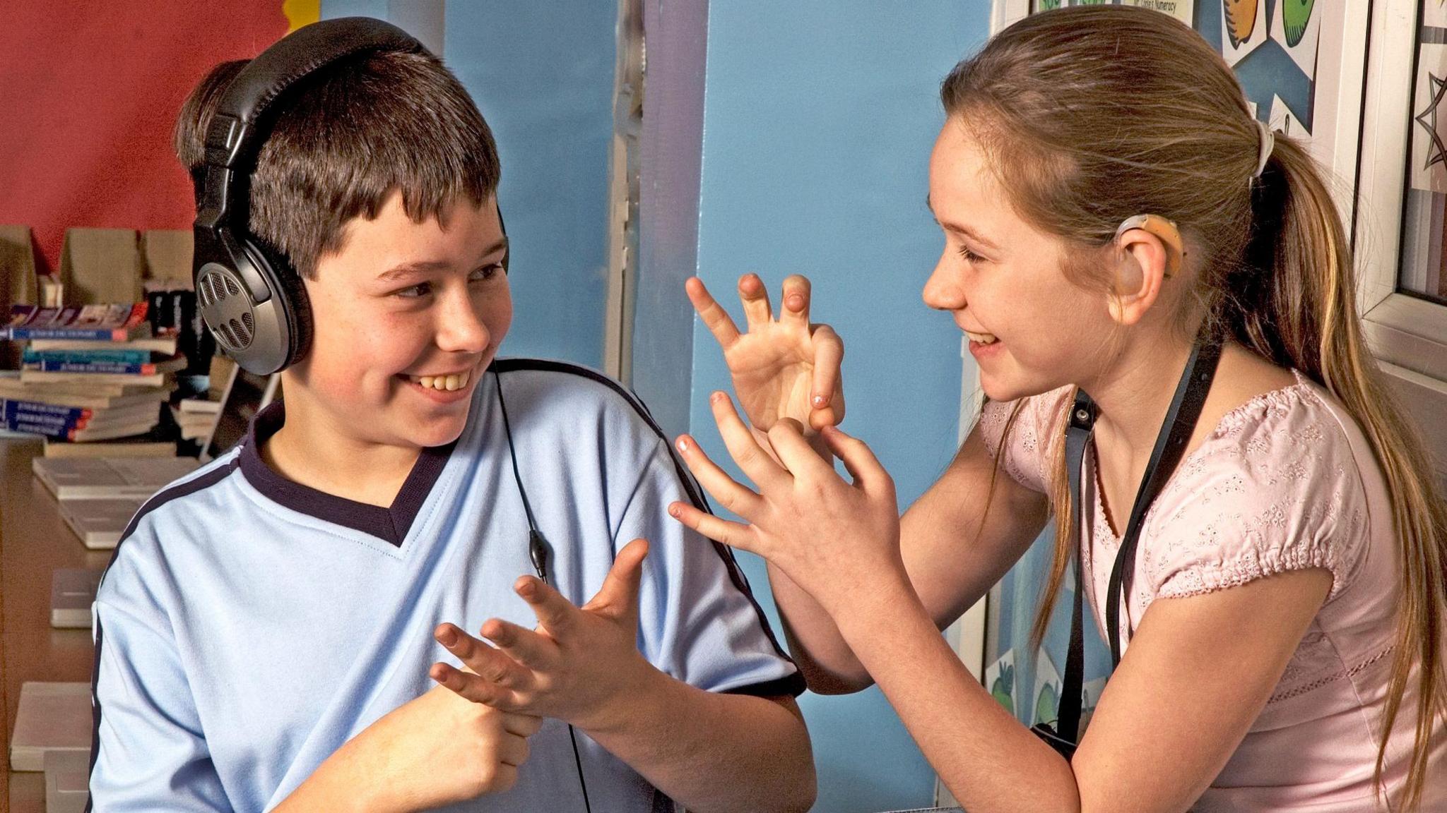 Deaf children with hearing aids in a school class room using sign language, a ten-year-old boy and a thirteen year-old girl communicate with their fingers and gesture, the boy wears head phones and the girl a hearing aid. The boy has a blue t-shirt and the girl is dressed in pink. The back ground are the walls of a school class room.