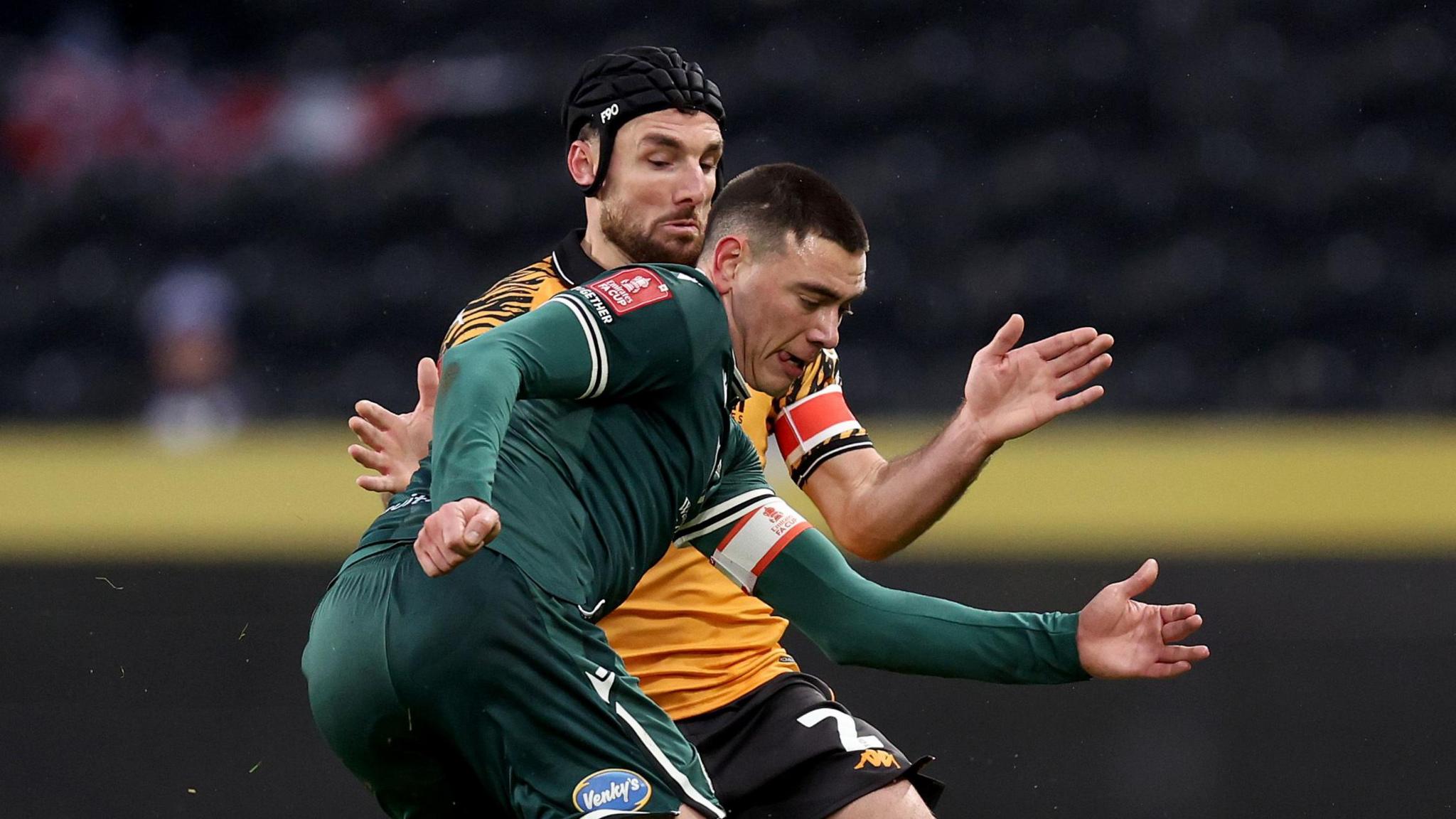 Blackburn captain Lewis Miller, in a dark green away kit, kicks the ball under pressure from Hull captain Lewie Coyle, who is wearing a scrum cap 