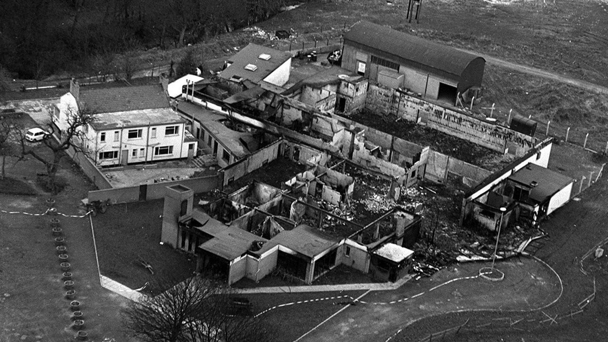 Black and white aerial image of the bombed La Mon hotel, the brick building is covered in black with the roof almost completely gone.  with parts with most of the building frame collapsed, debris lies on the floor.