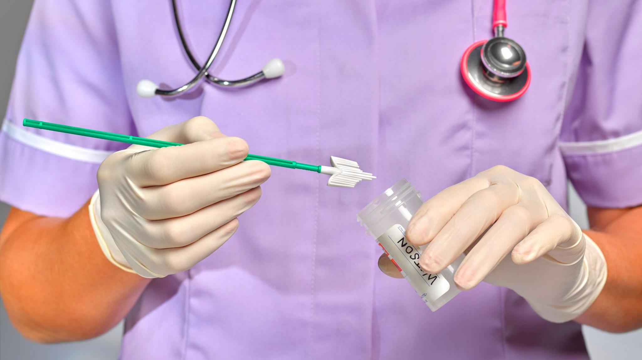 A nurse in a purple uniform places a sample, on the end of a green stick, into a clear and colourless tube. She has a stethoscope around her neck and wear white gloves.  