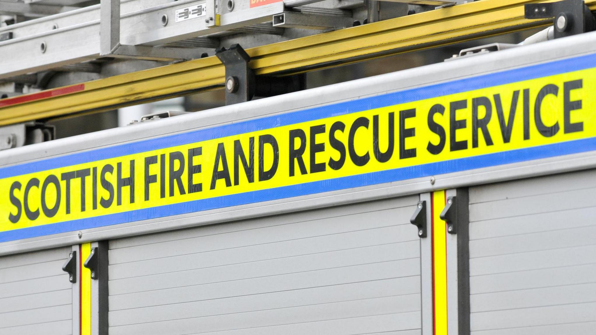 A close up of the side of a fire engine. There are ladders on the roof of the vehicle with the words "Scottish Fire and Rescue Service" written on it in black letters on a yellow background.