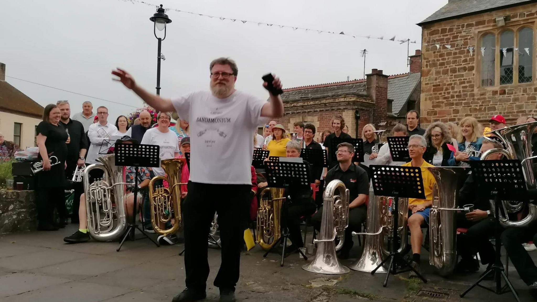 A picture of Tim stood in front of a brass band wearing a white shirt. He has a grey beard, holding a black phone and is wearing glasses. He is smiling at the camera.