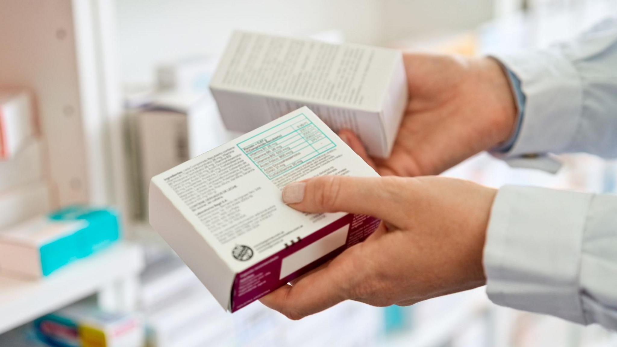 A person wearing a white lab coat holding two white boxes of medicine in each hand.