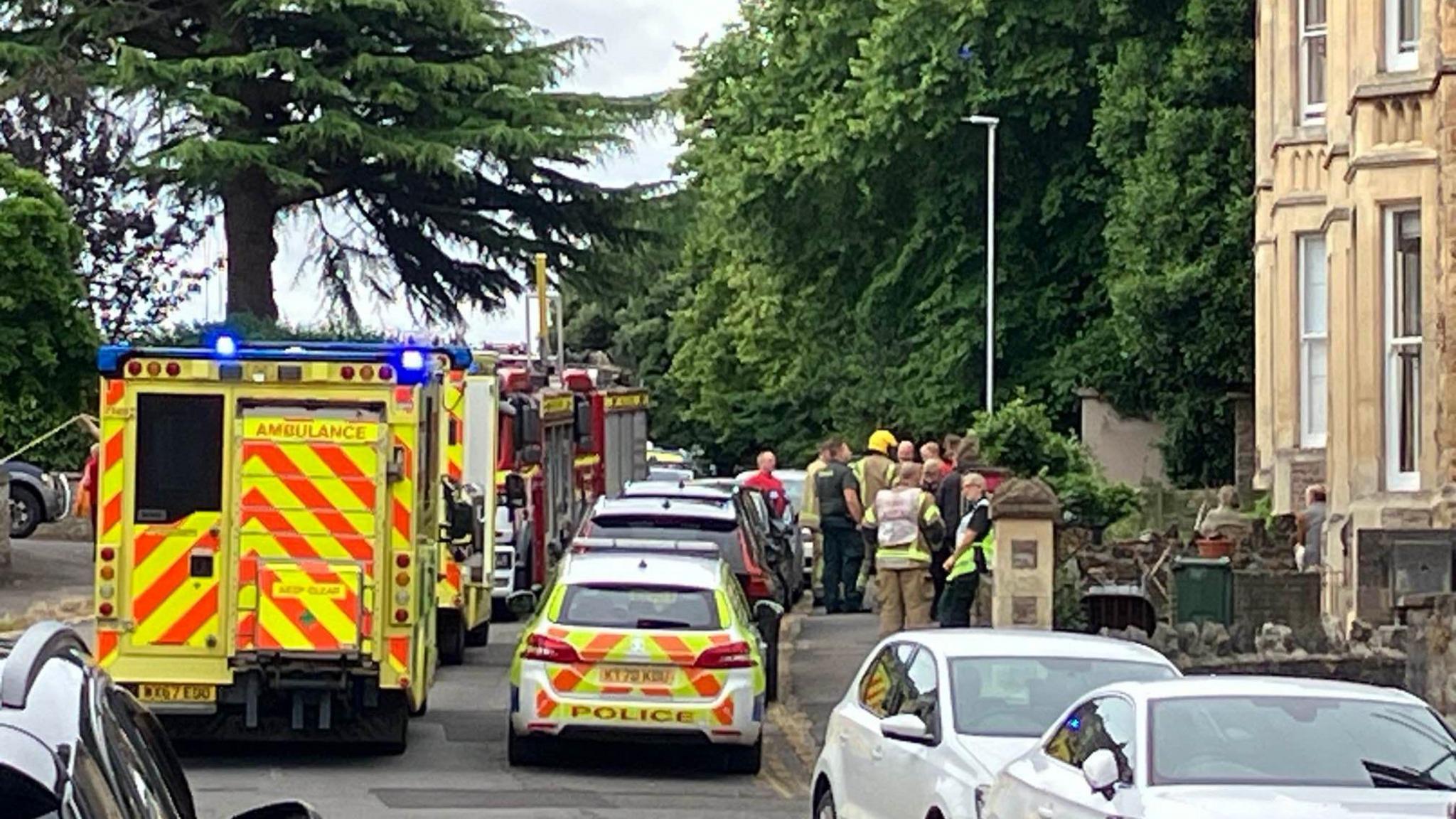 The back of a police car, ambulance and fire truck parked on Victoria Road. There are paramedics standing outside a property and looking up at the windows