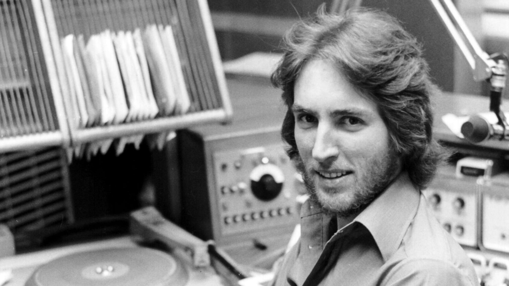 Black and white image of Johnnie Walker pictured in a BBC radio studio in 1972. He has a feathered haircut and beard and is wearing an open-necked shirt.