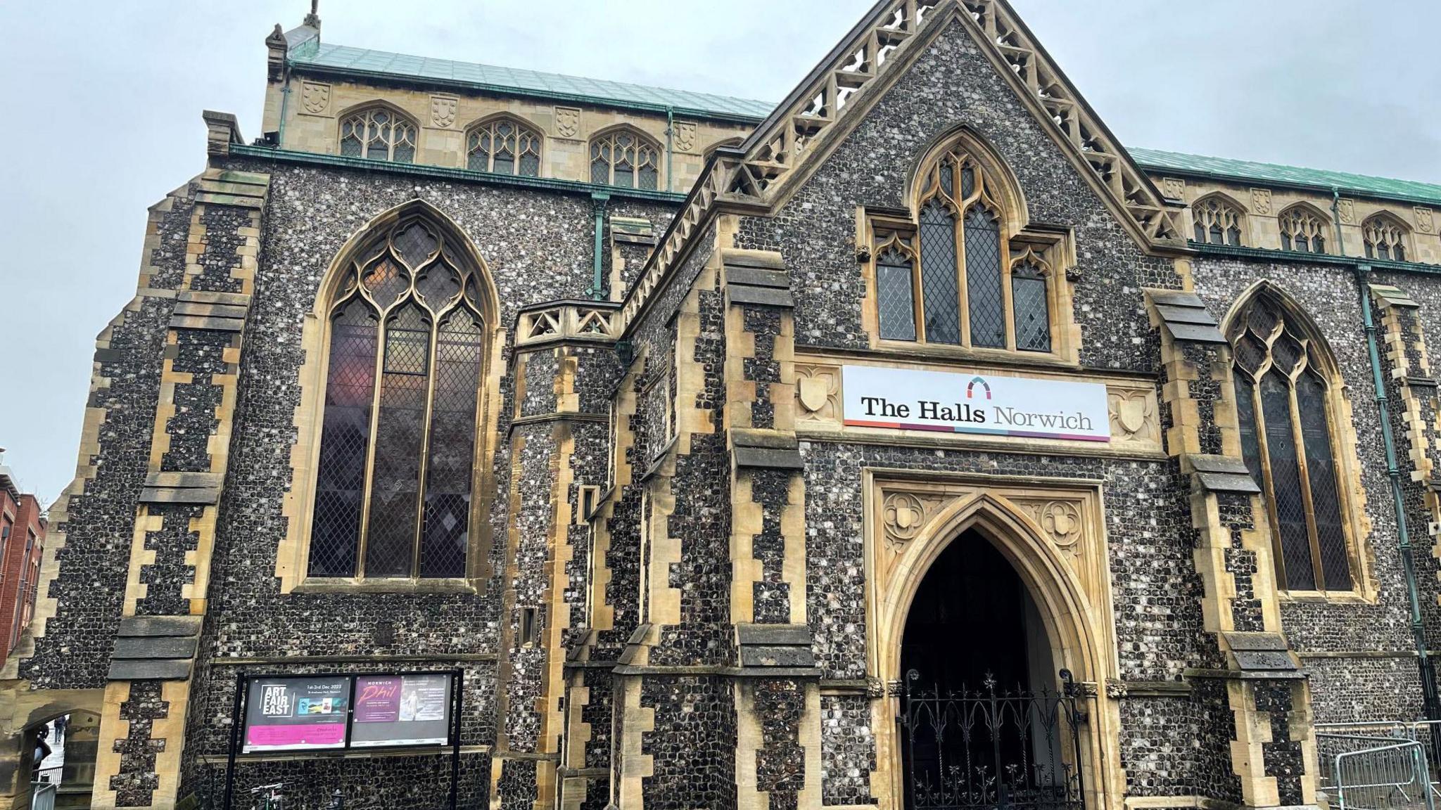 The exterior of a former church which contains the Blackfriars and St Andrew's Halls in Norwich city centre. It has a flint exterior, arched windows and an arched entrance. Above the main door is a sign saying "The Halls Norwich".