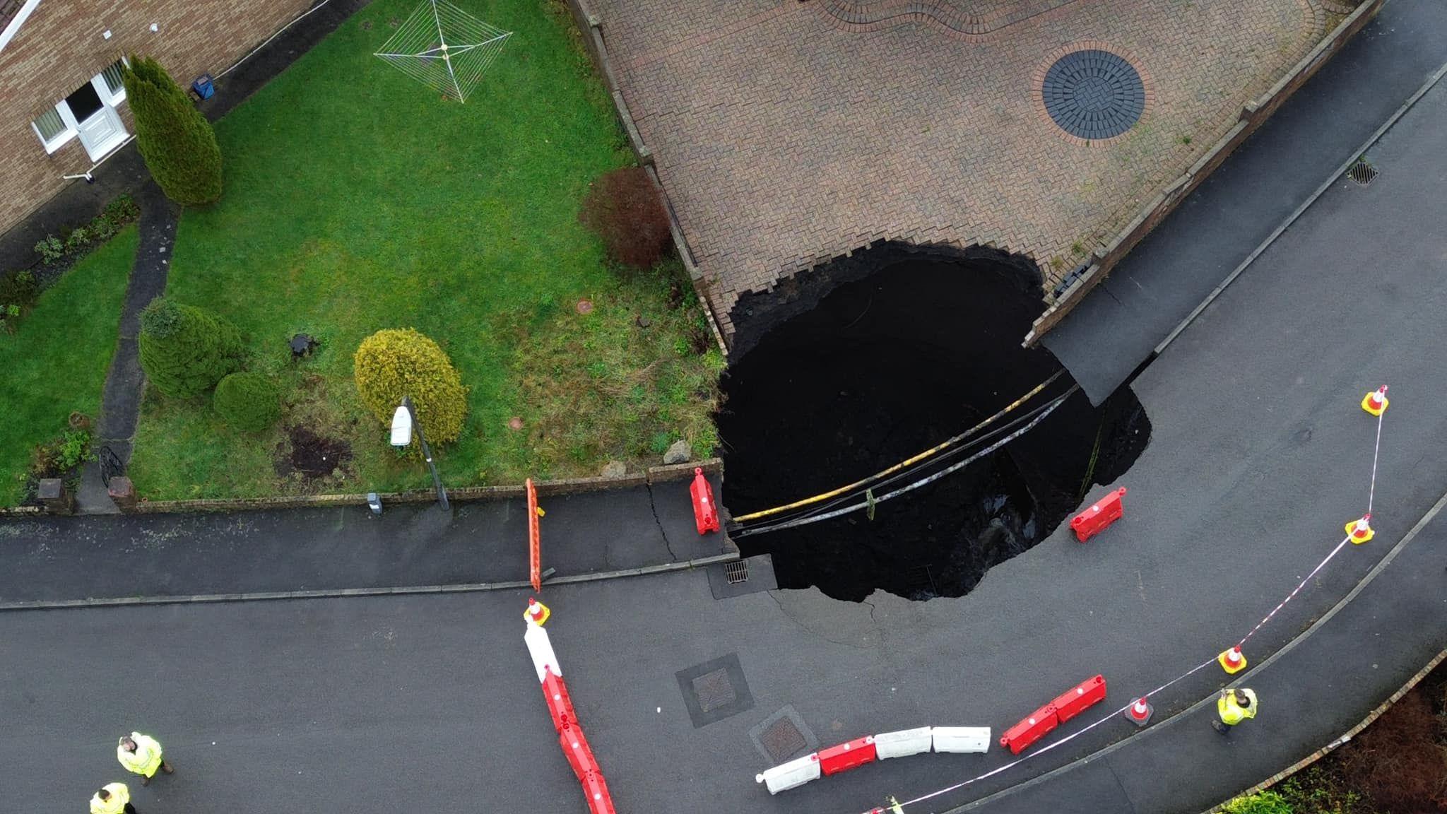 An aerial image of the sinkhole with people's drives and gardens, plastic barriers and emergency services around it